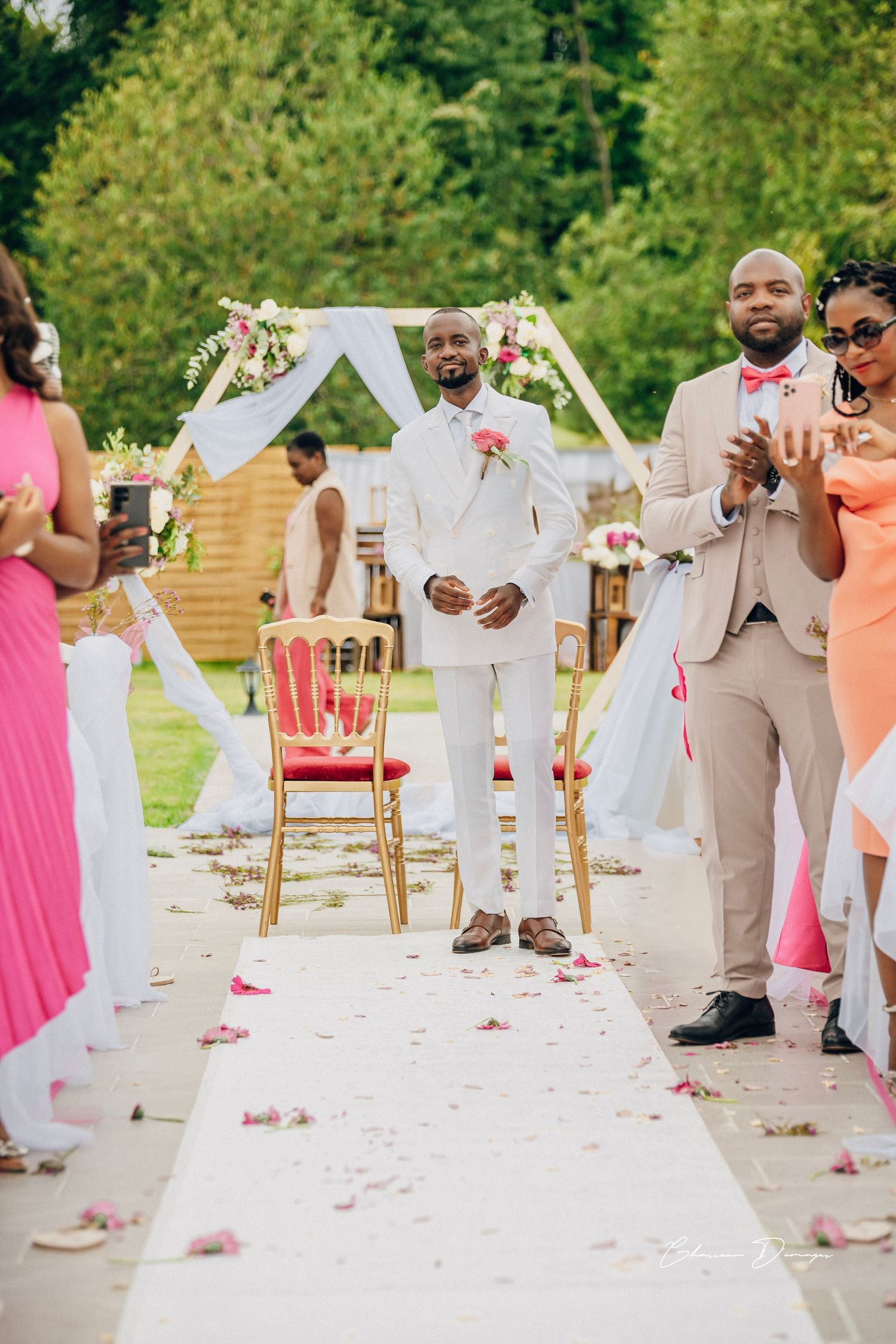 Wedding ceremony setup with white arch, pink flowers, and white aisle runner outdoors.