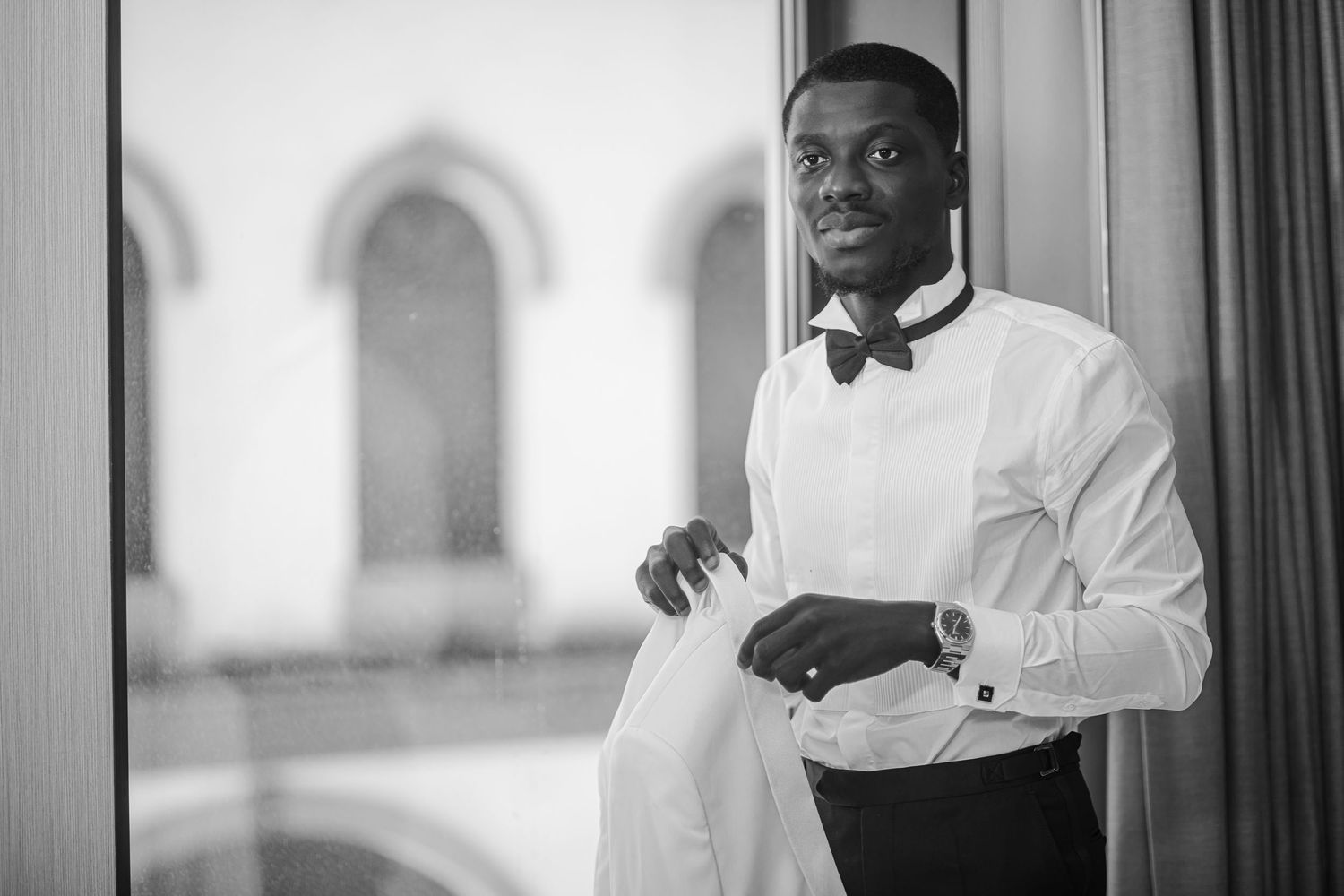 Black and white portrait of a person in formal attire with a bow tie standing near a window.