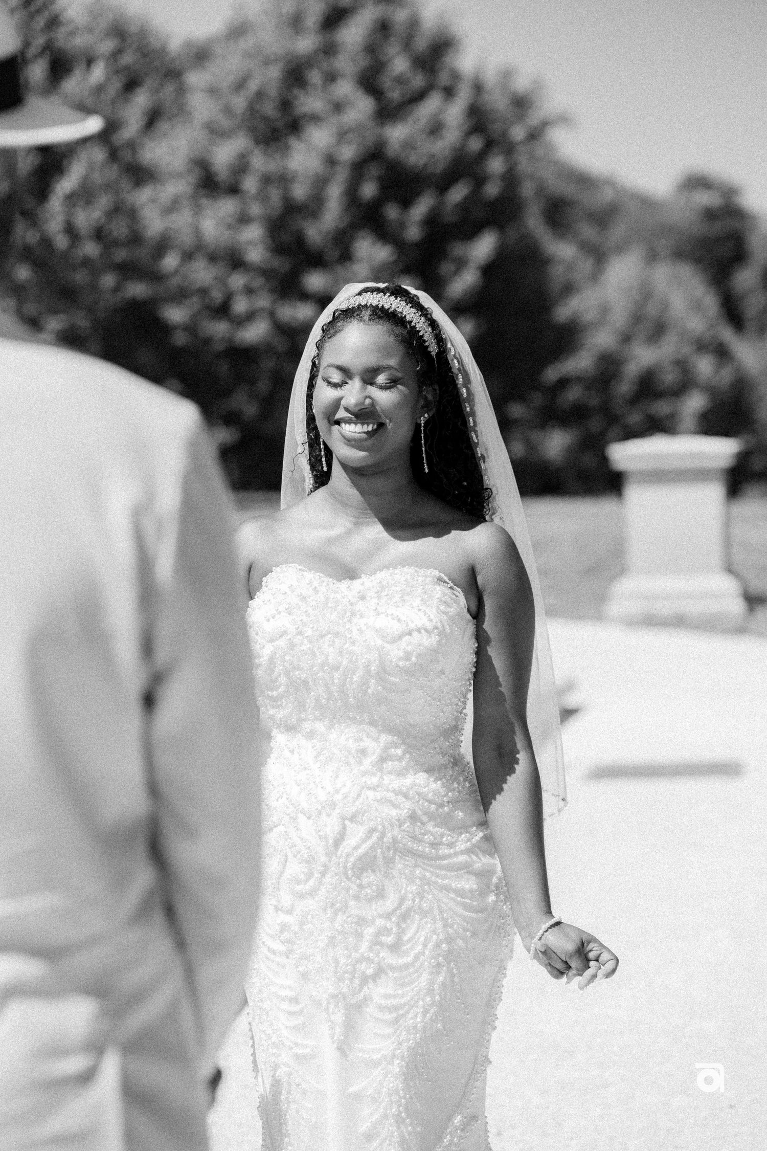 A radiant bride in a fitted lace wedding dress with veil smiles joyfully during an outdoor ceremony.