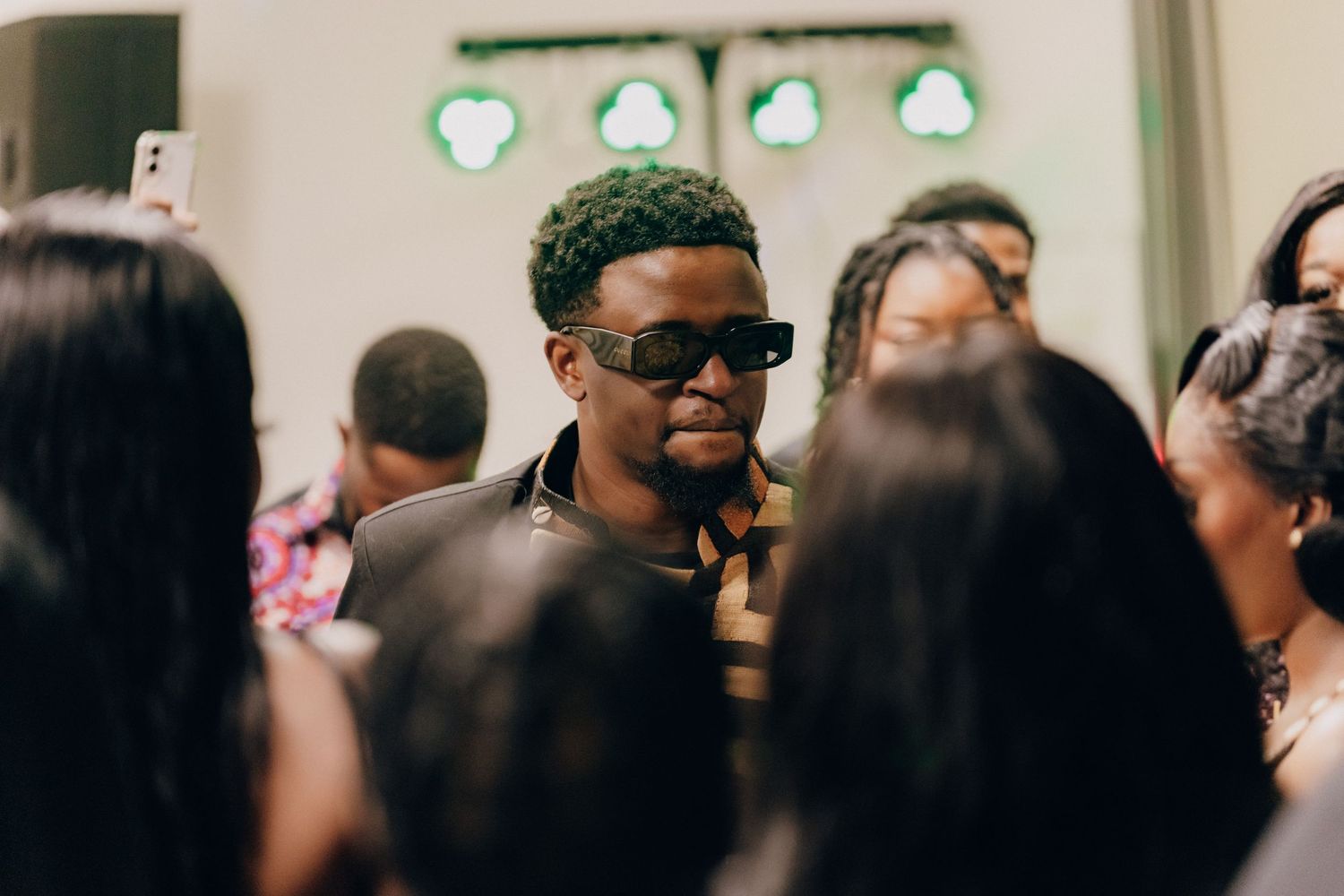 A stylishly dressed person wearing sunglasses amid a crowd at an indoor gathering.