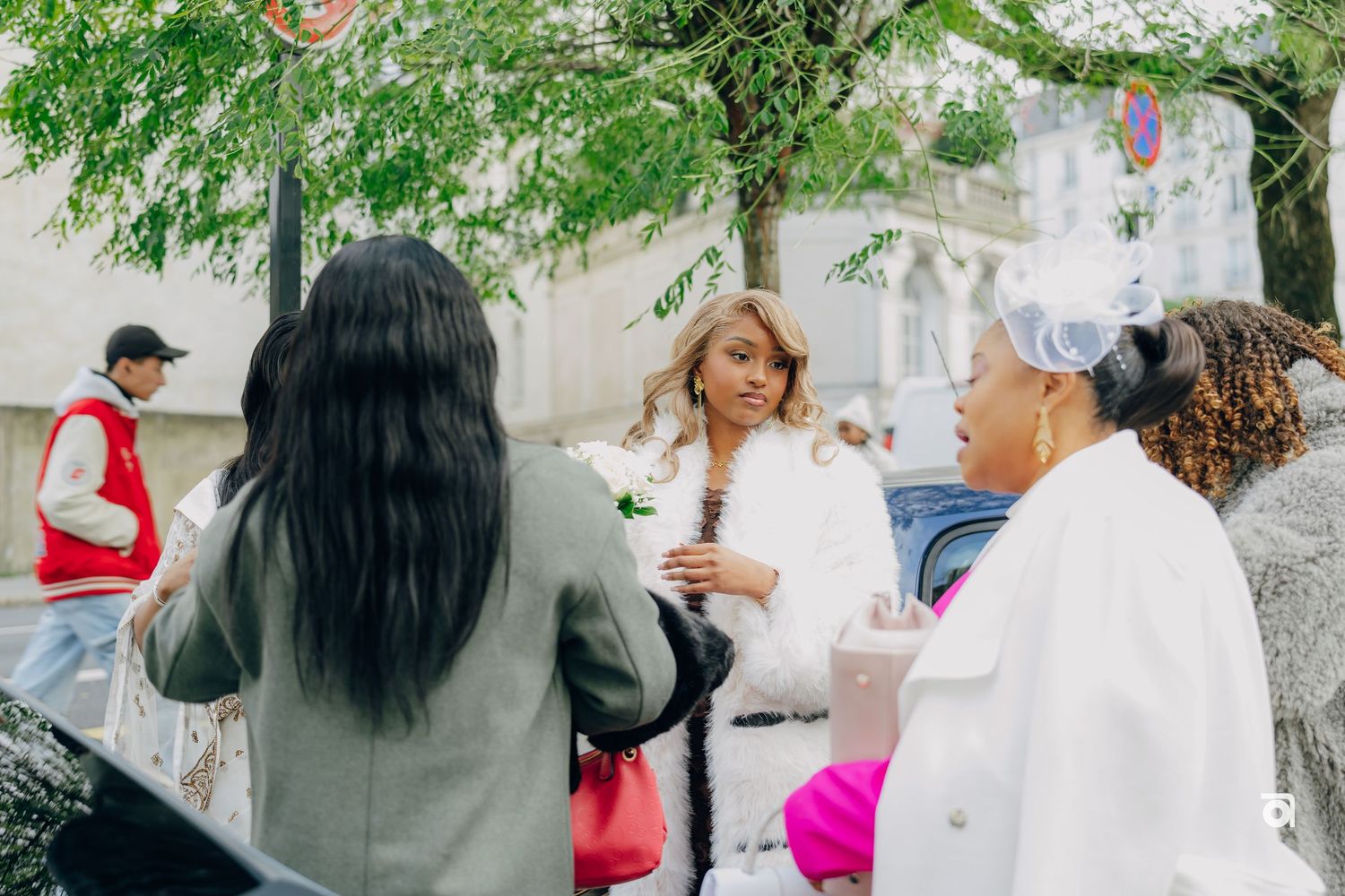 A group of people gathered outdoors having a conversation near leafy trees and buildings on a sunny day.