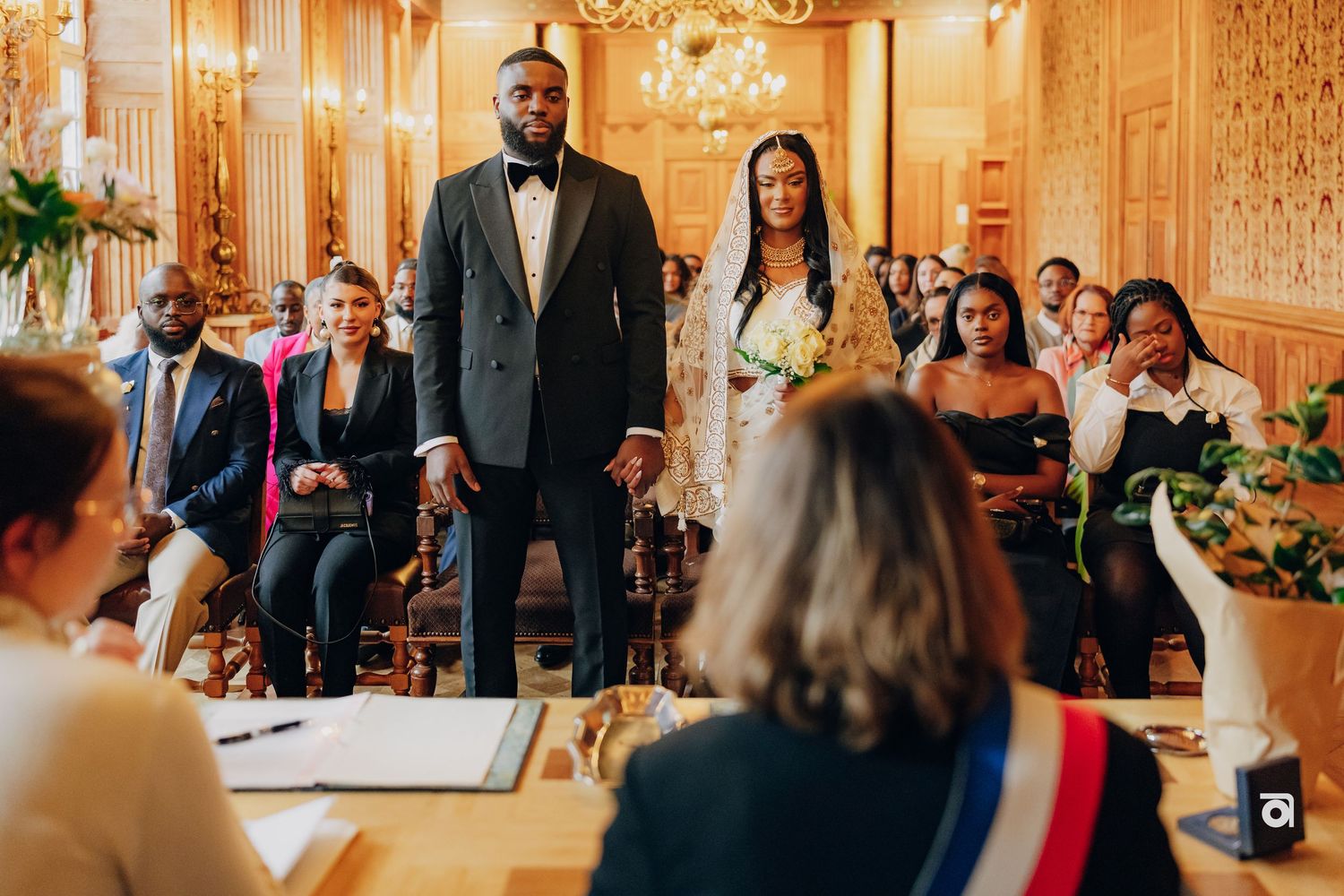 Wedding ceremony taking place in an ornate golden room with chandeliers and wooden details.