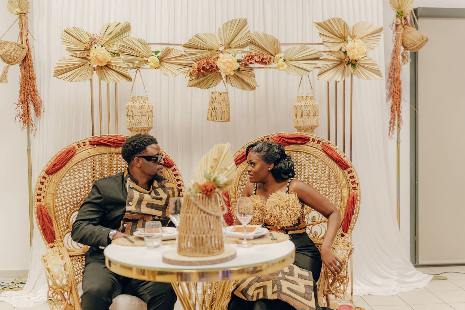 Two people enjoy hot beverages while seated at an elegant round table with golden decorative elements above.