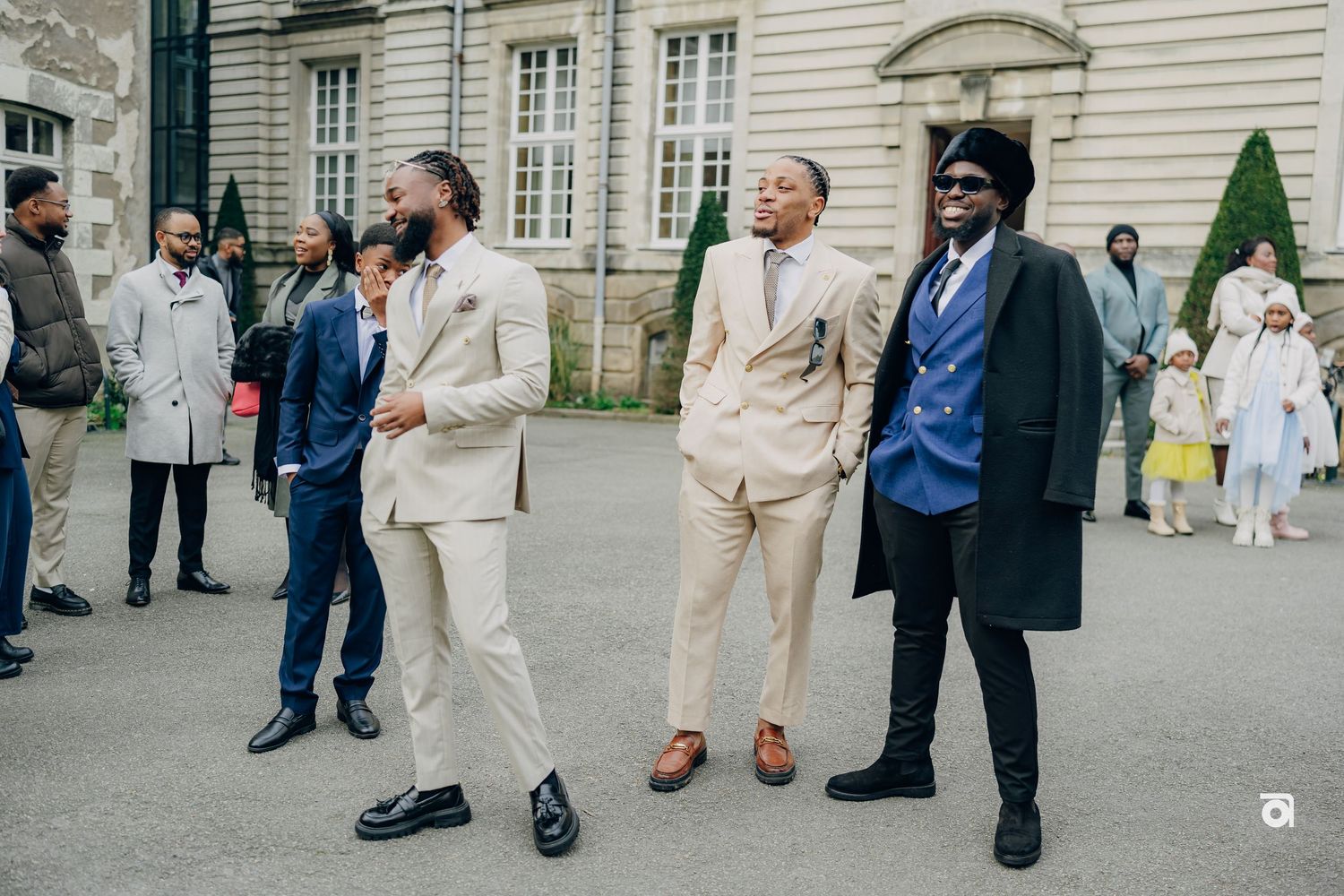 A group of well-dressed men in suits and formal attire gather outside an elegant building with classical architecture.