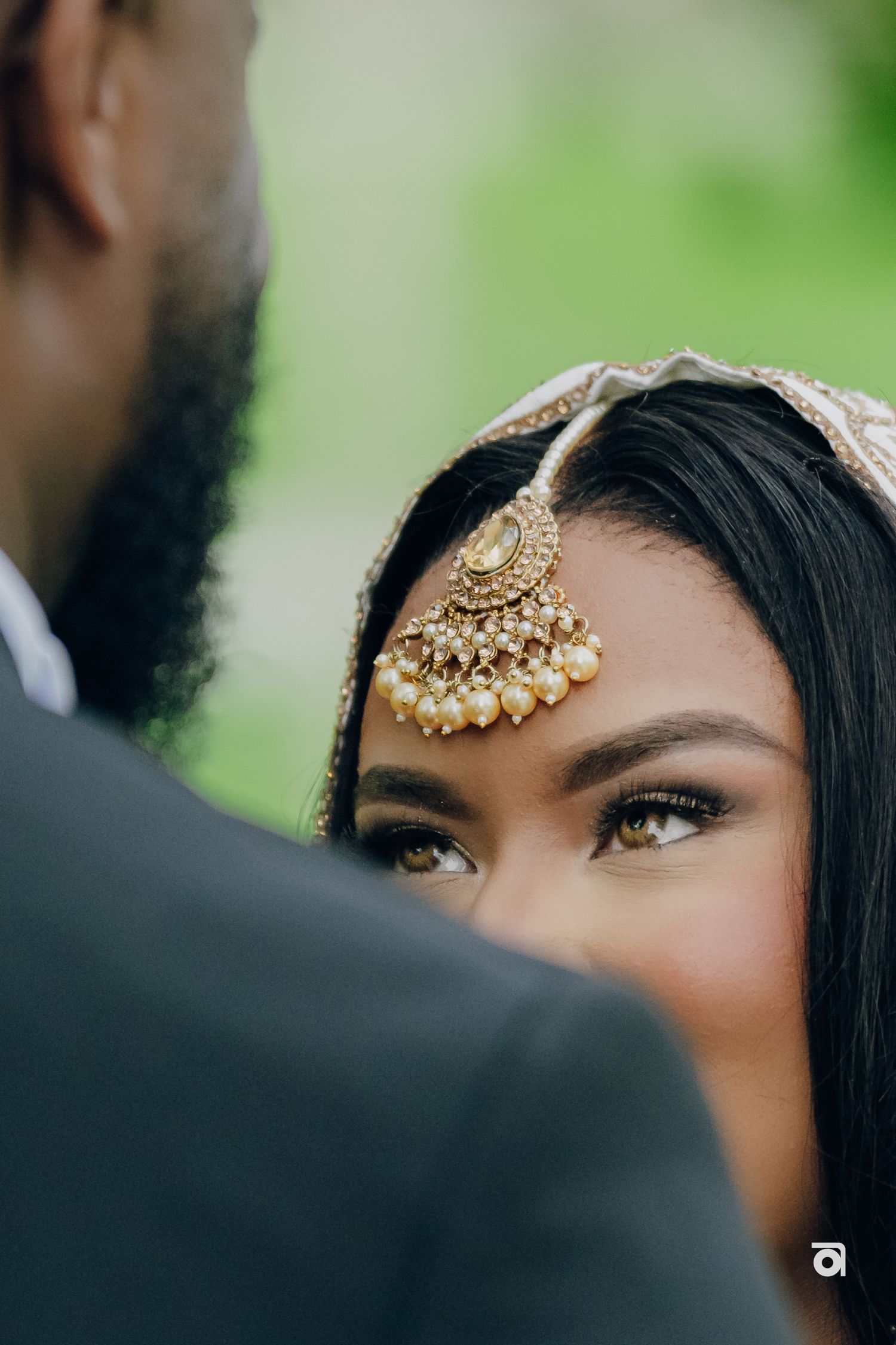 Close-up side view of someone wearing an ornate gold tikka headpiece with pearls during traditional South Asian wedding.