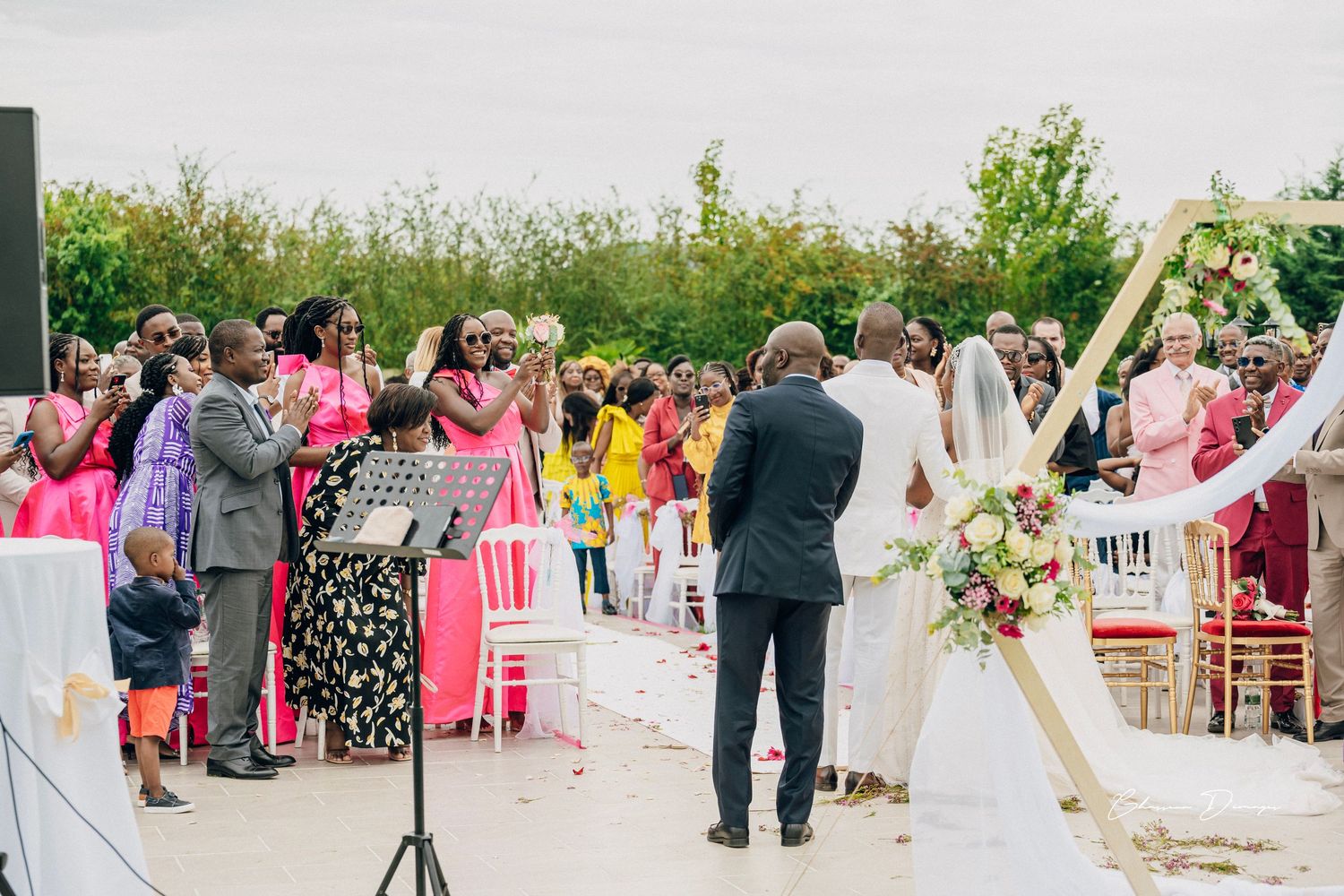 Wedding guests celebrate as newlyweds exit their outdoor ceremony under a decorated arch.