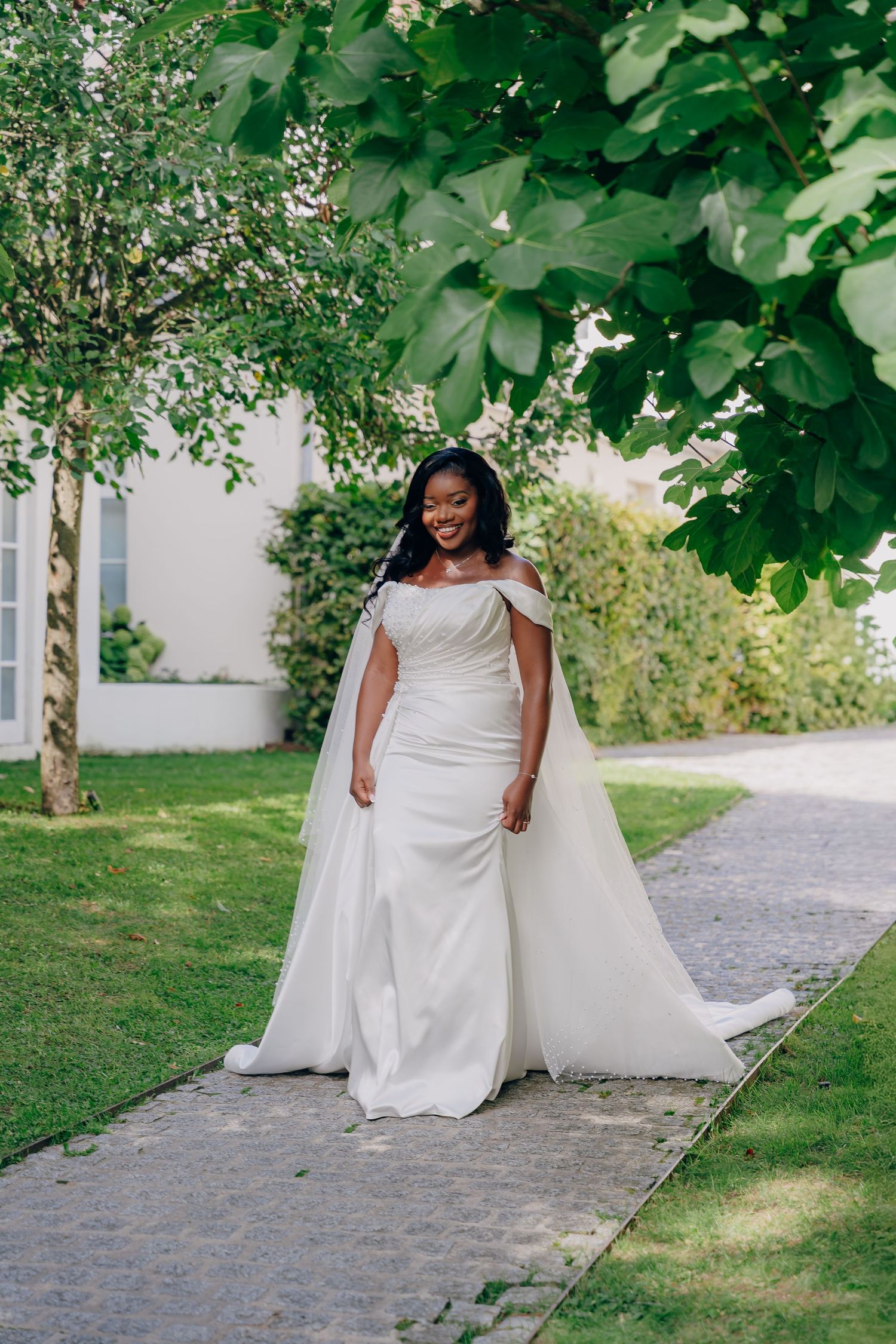 A wedding dress flows gracefully along a garden path lined with lush green trees and foliage.