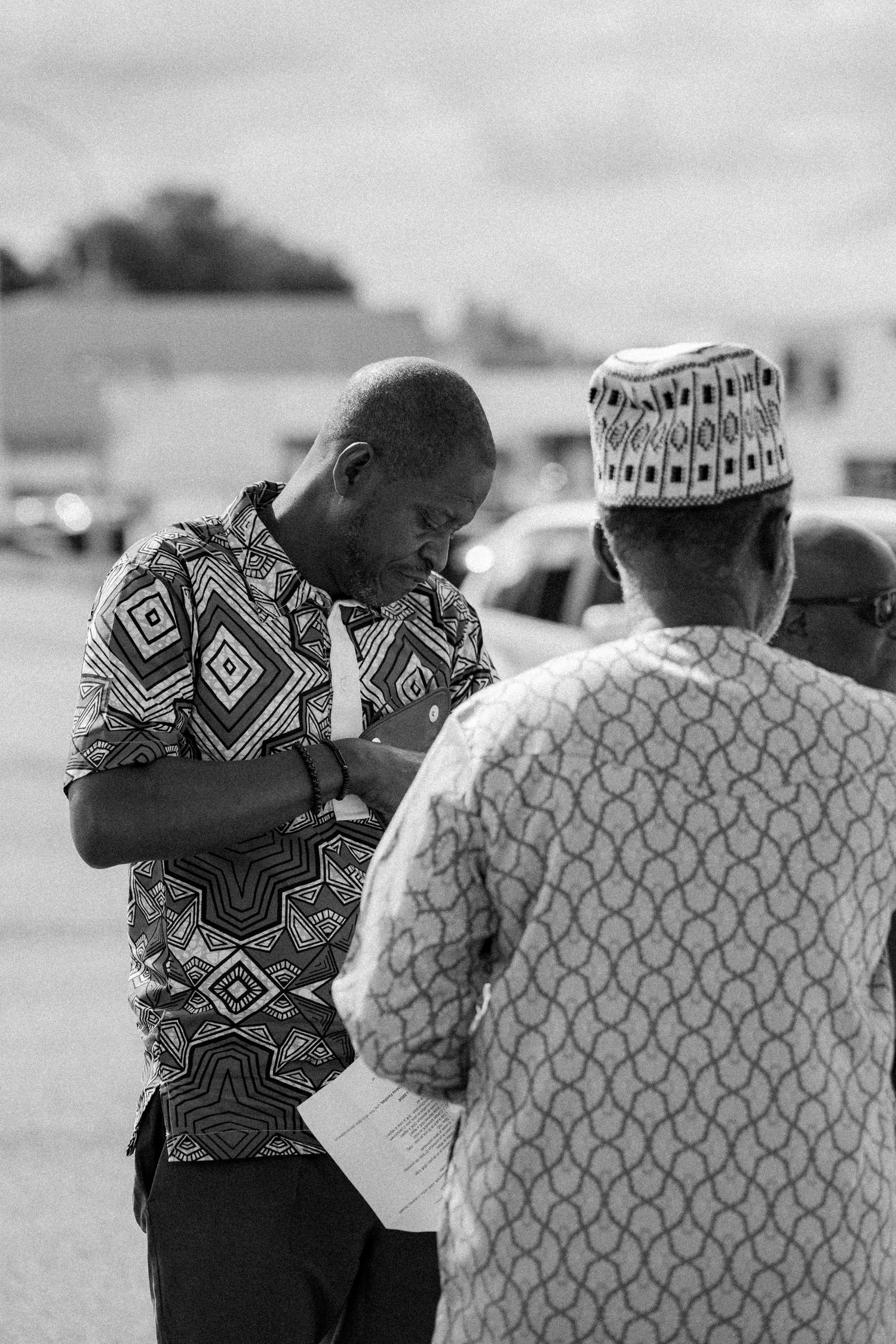 Two men in patterned shirts stand in conversation outdoors, with one wearing traditional African attire.