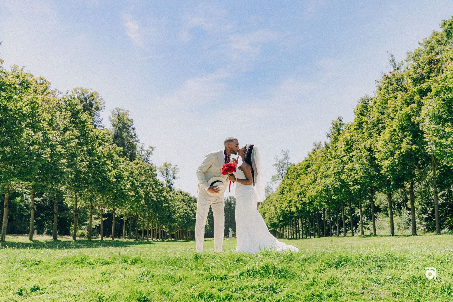 A couple embraces in a scenic park setting with green trees lining a pathway on a beautiful summer day.