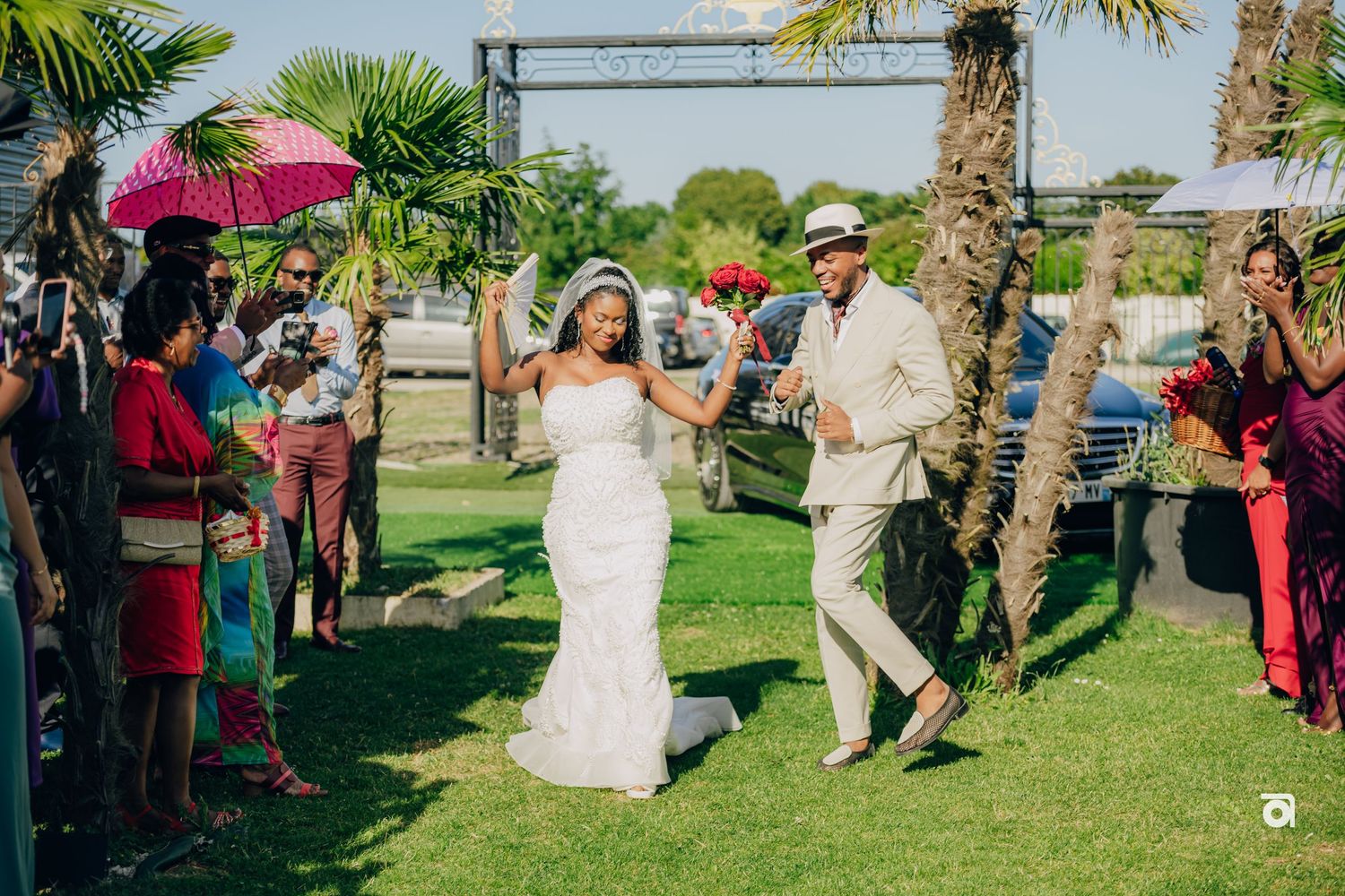 A wedding celebration on a sunny lawn with attendees holding pink parasols while a couple walks down the grassy aisle.