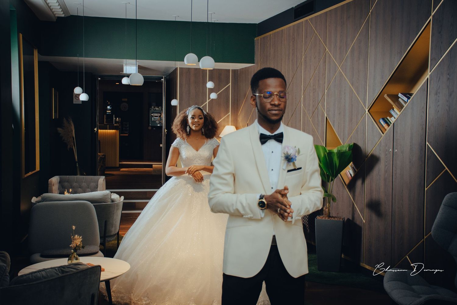A couple on their wedding day pose in an elegant indoor venue with the groom in a white tuxedo jacket.