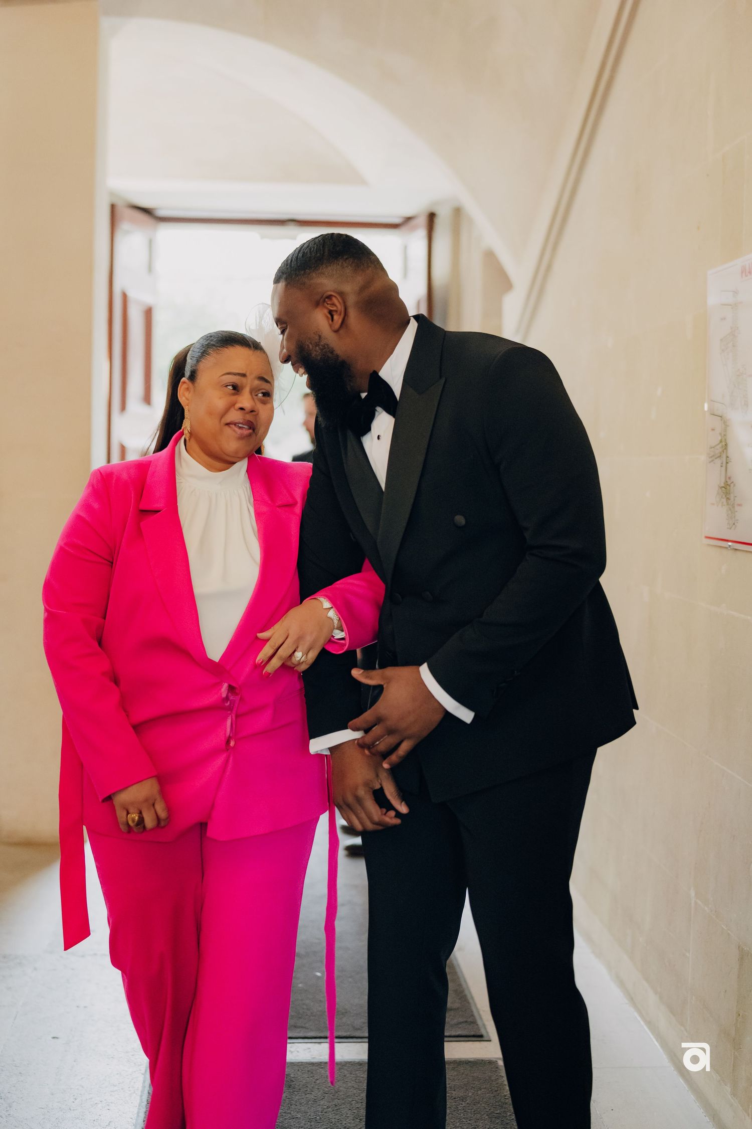 Two people share a joyful moment in a hallway, one wearing a bright pink suit and another in a black tuxedo.