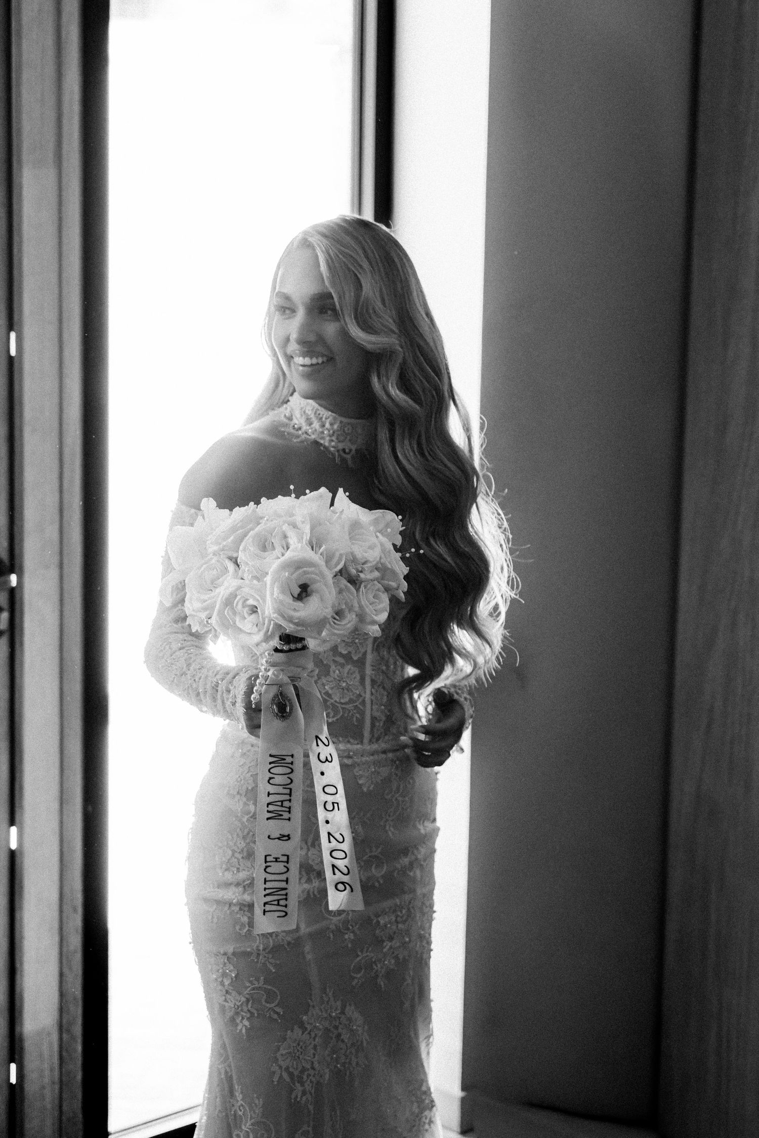Smiling bride in lace off-shoulder gown holds white floral bouquet with ribbon, black and white photo.