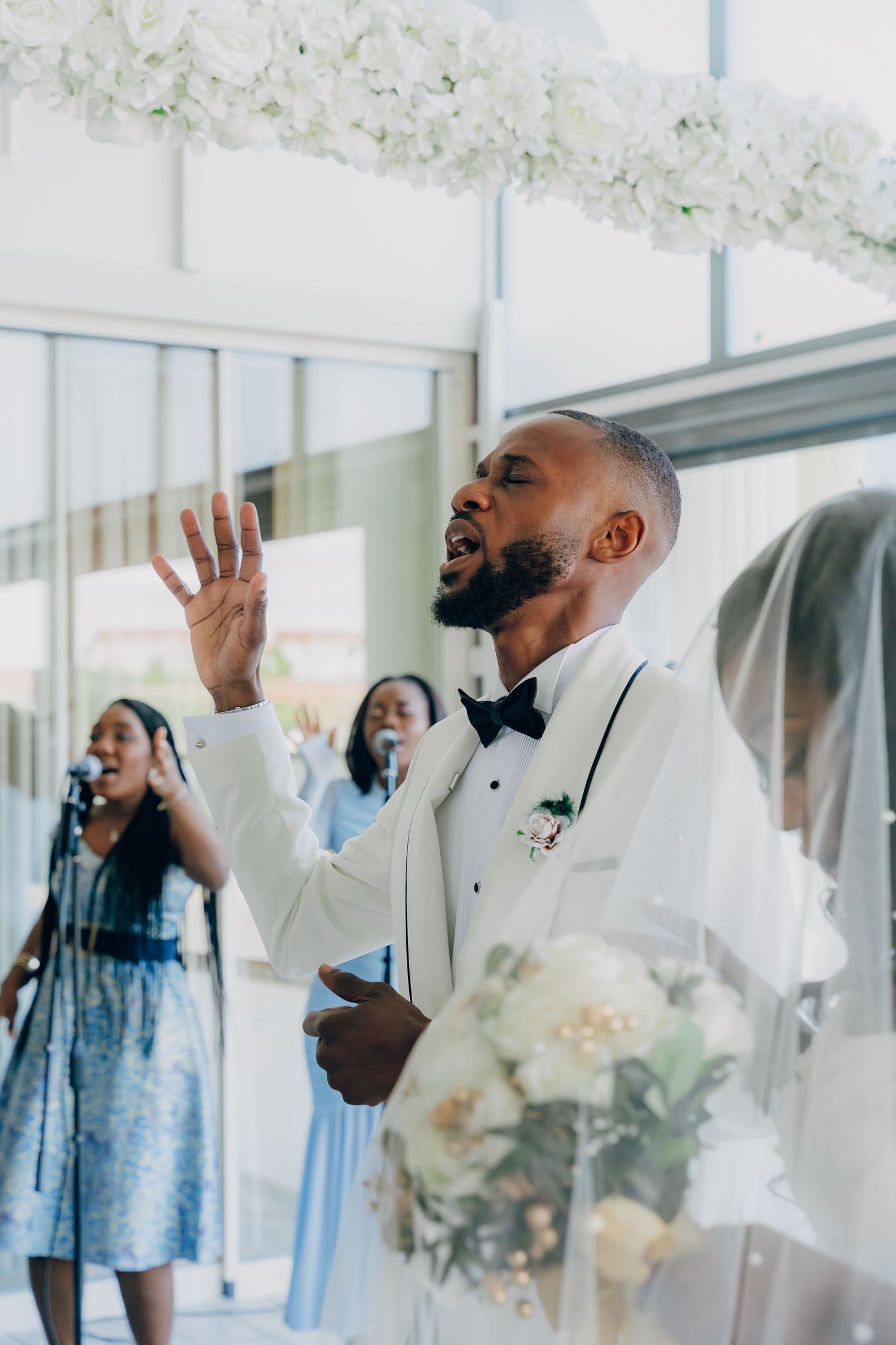 Wedding guest in white tuxedo raises hand emotionally during ceremony under floral decorations.
