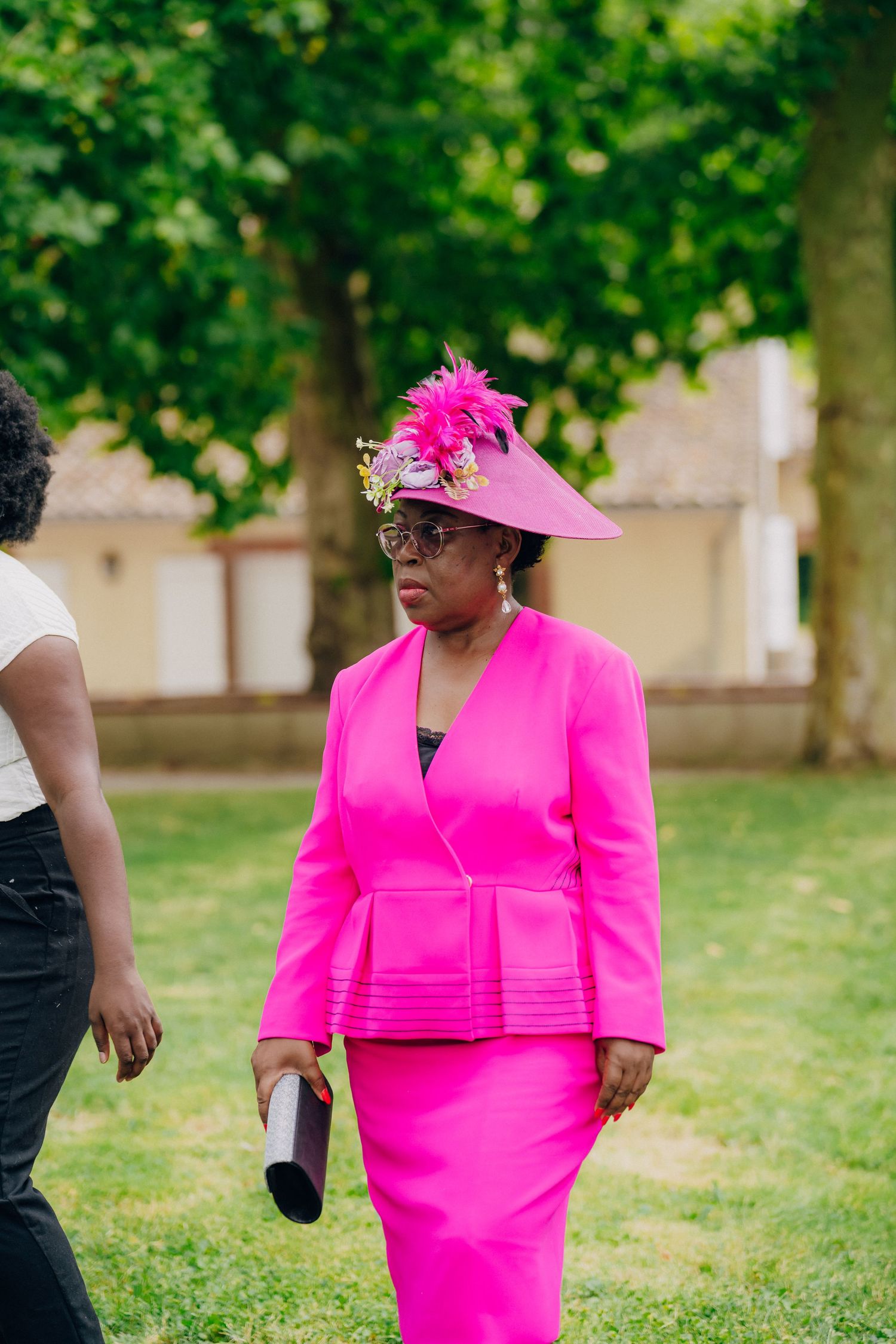 A person wearing a vibrant hot pink dress and matching hat stands on a grassy lawn during an outdoor event.