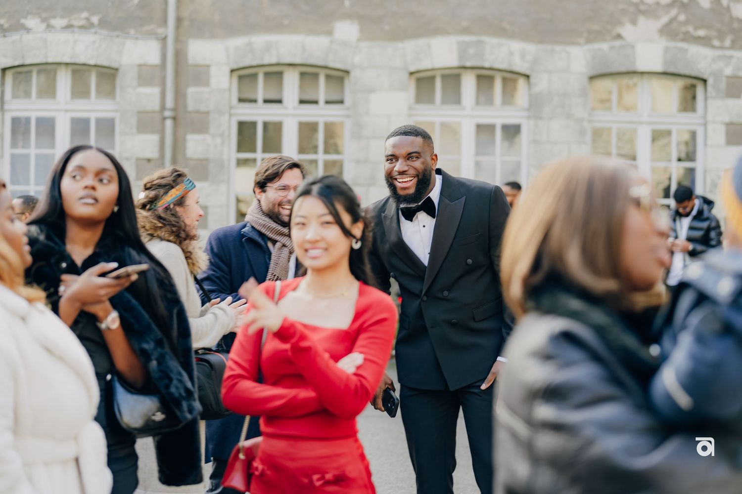 A group of wedding guests gather outside a building with arched windows during a celebration event.