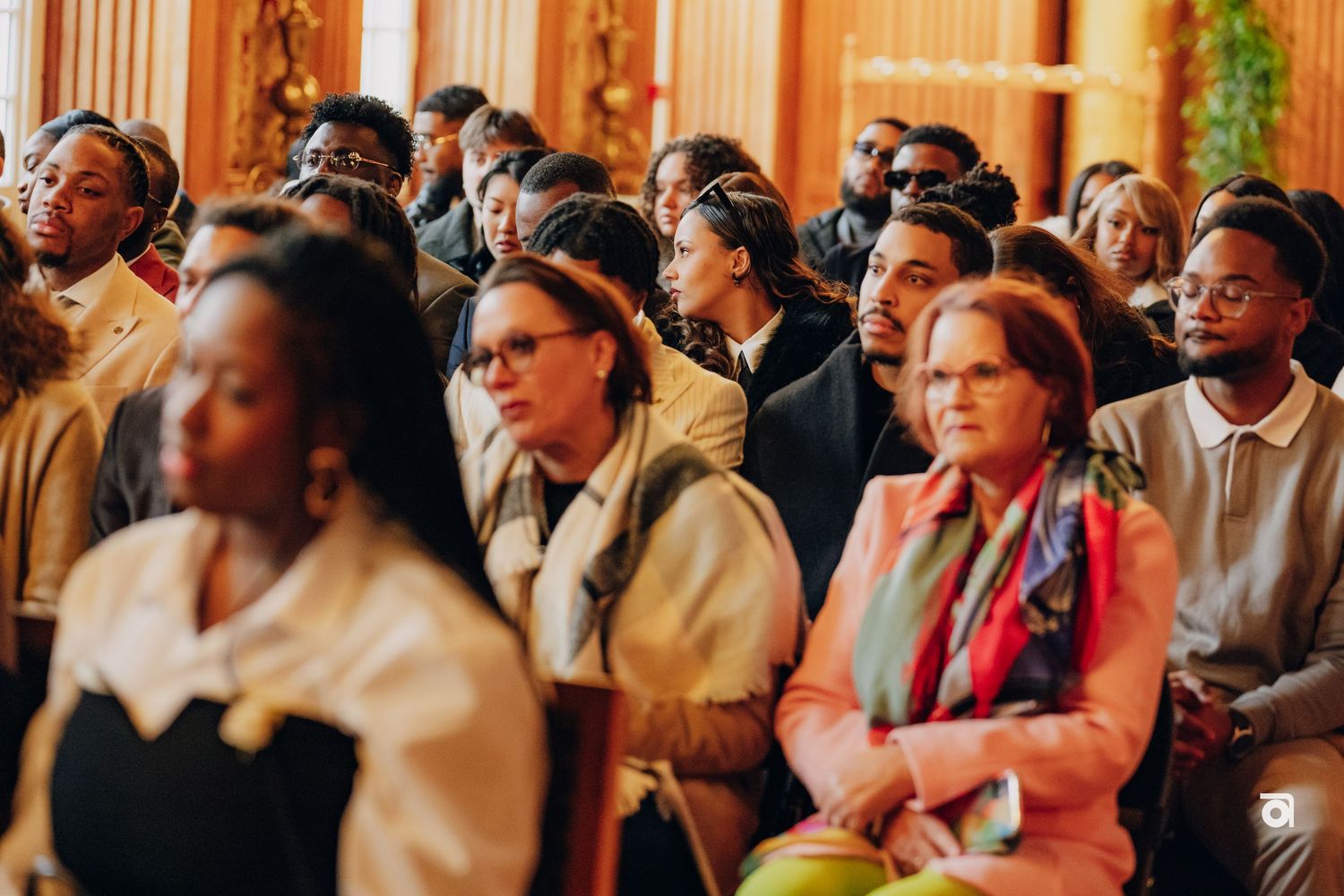 An attentive audience seated in formal dress at an elegant venue with ornate decor.