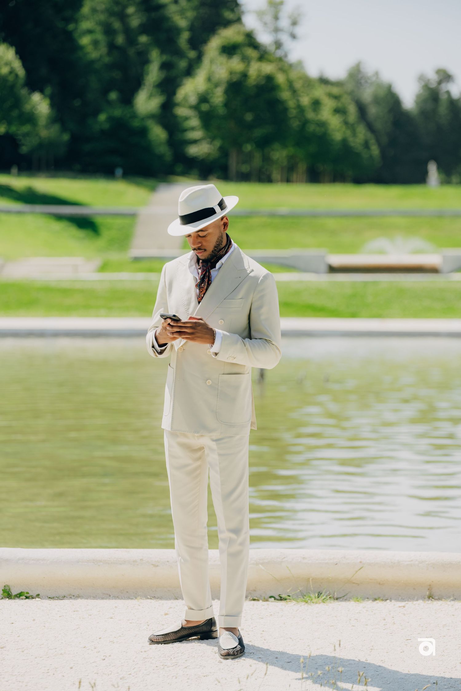 A person in a crisp white suit and panama hat checks their phone beside a serene pond on a sunny day.