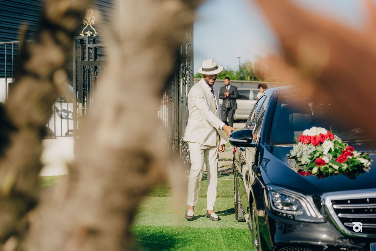 A person wearing a light colored suit stands next to a black car with pink and white flowers on top at an outdoor event.