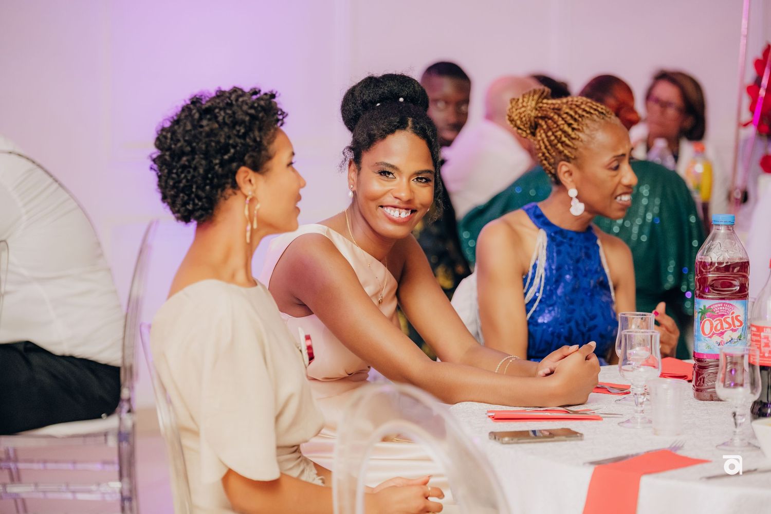 Three friends enjoying drinks and conversation at a pink-themed event, smiling and chatting at a table.