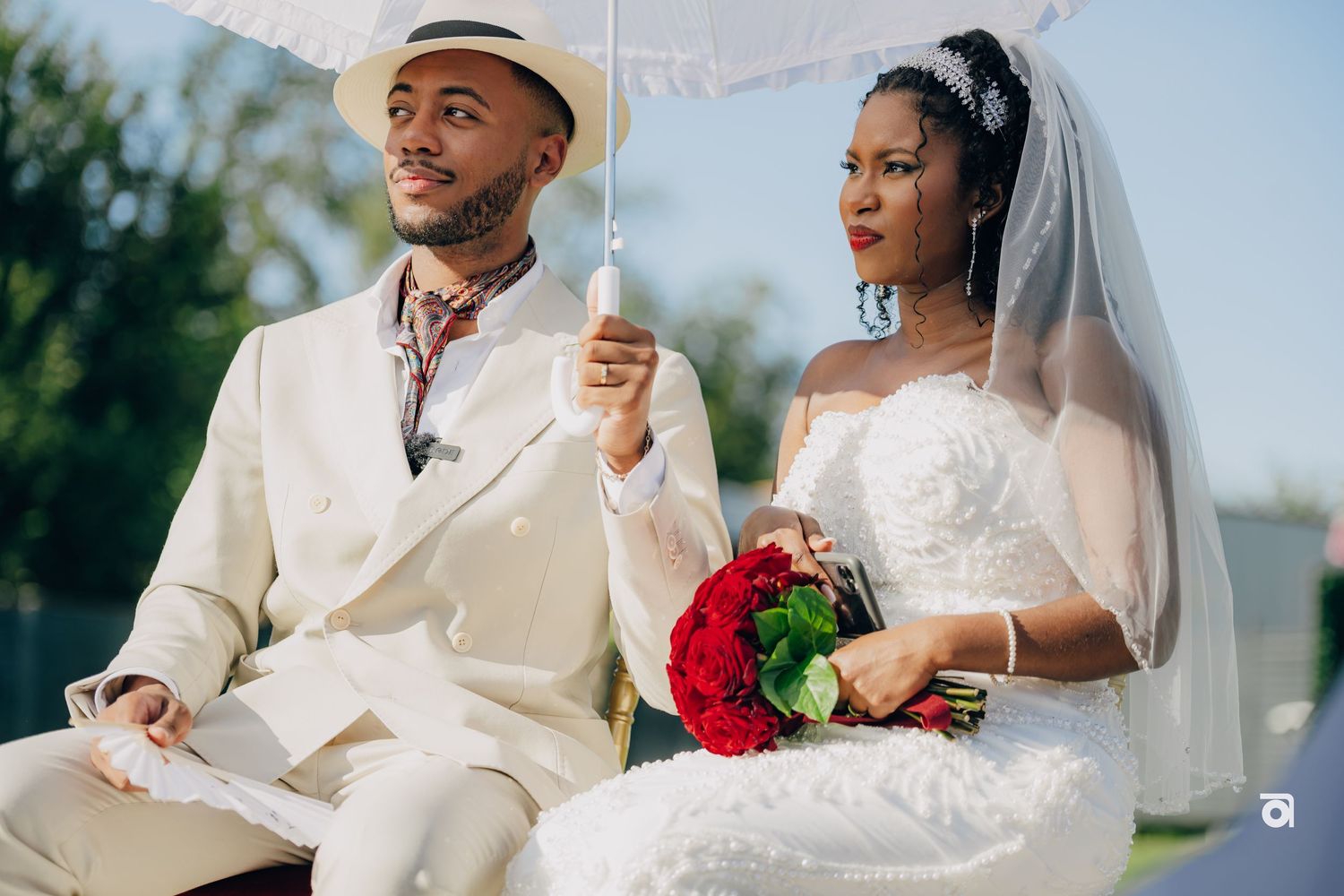 A bride in white gown holds red roses while seated next to groom wearing cream suit during outdoor ceremony.