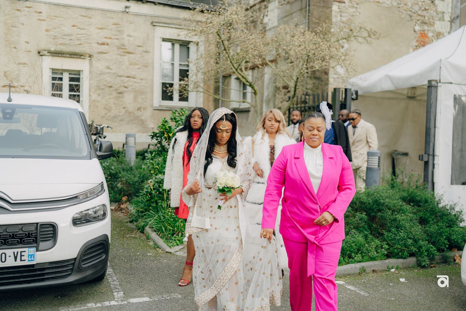 A couple walks outdoors near a stone building, with the person on right wearing a vibrant pink suit.