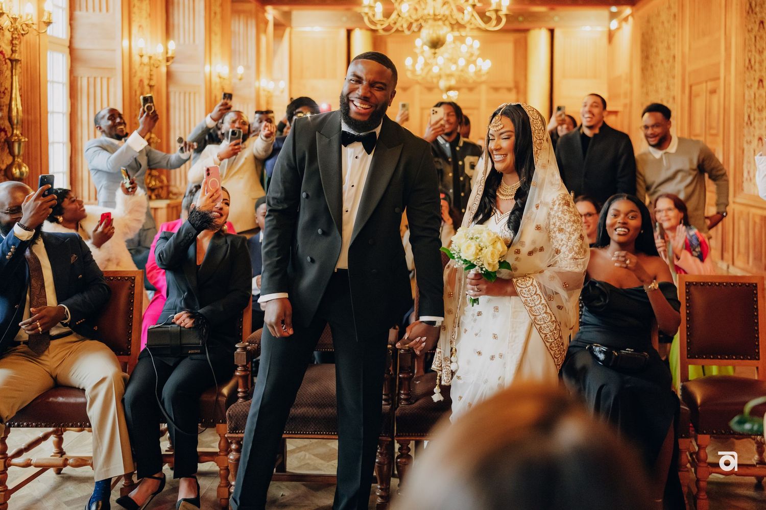 A celebratory scene in a golden-lit reception room with guests cheering the newlyweds.