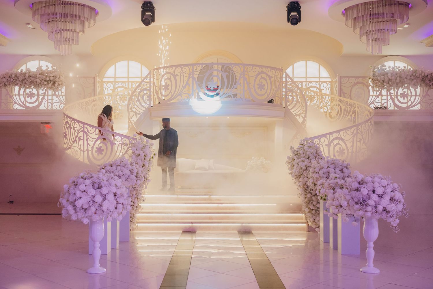 Luxurious wedding venue with arched windows, chandeliers and white floral arrangements lining the aisle.