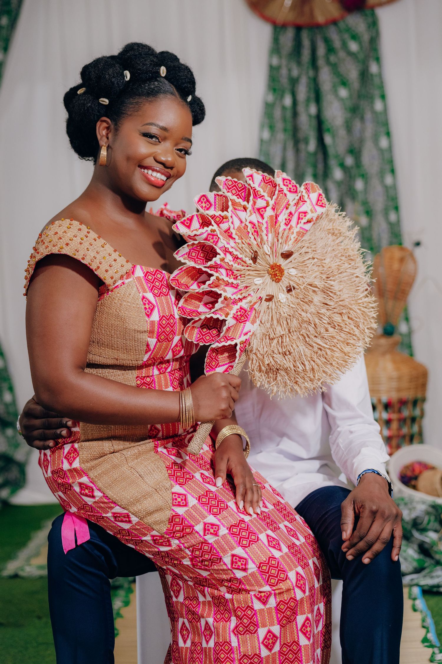 People in traditional pink and beige attire holding decorative woven fans during a celebration.