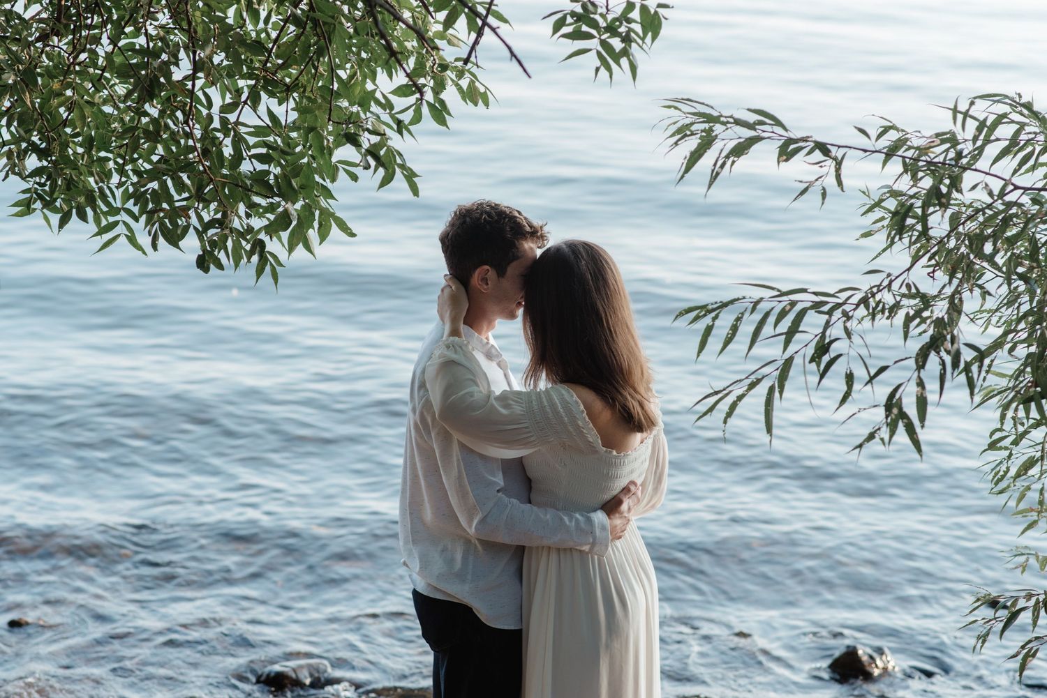 A couple embraces at the waterfront beneath willow branches hanging over a serene lake on a tranquil day.