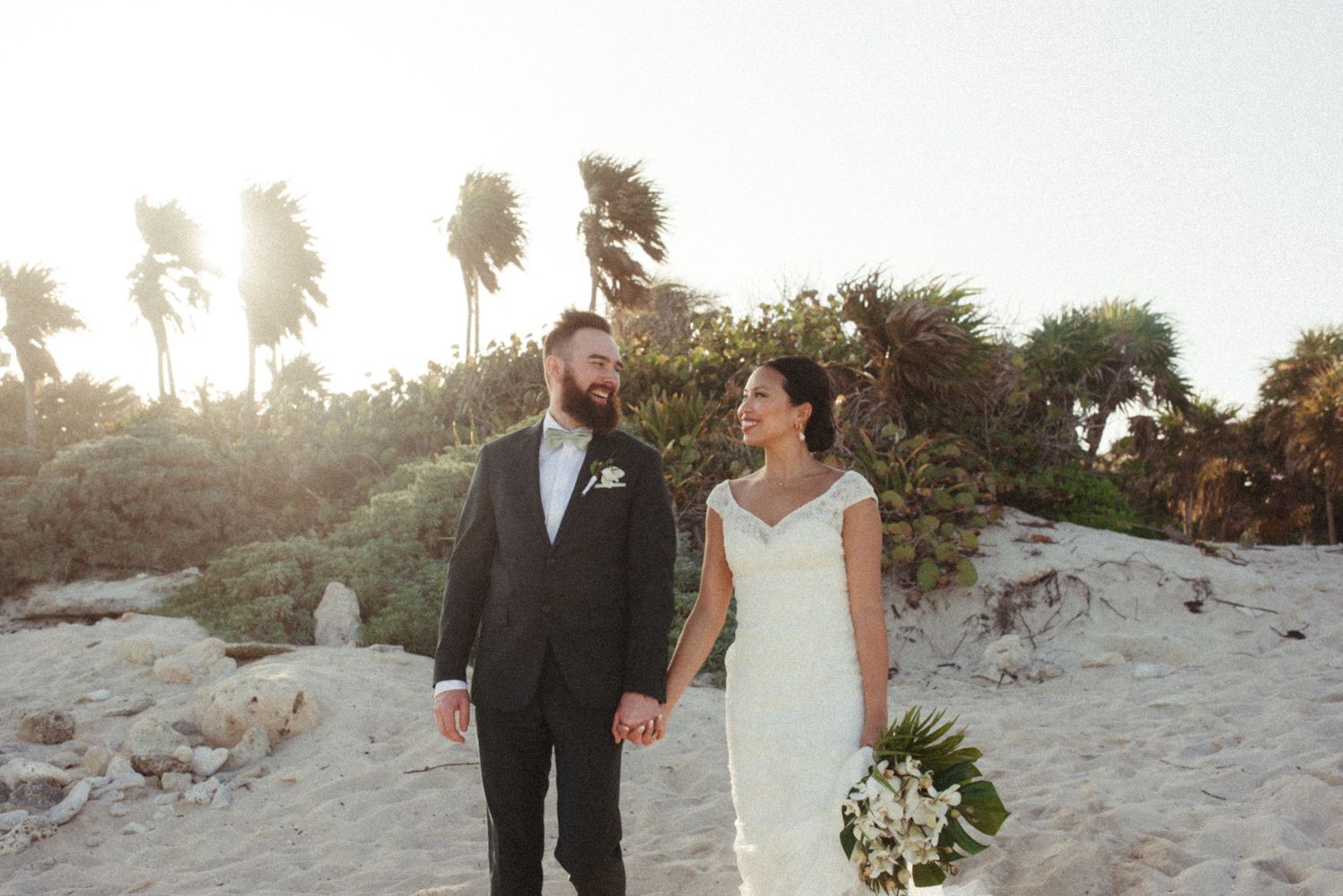 A couple walking together on a sandy beach at sunset with palm trees in the background.