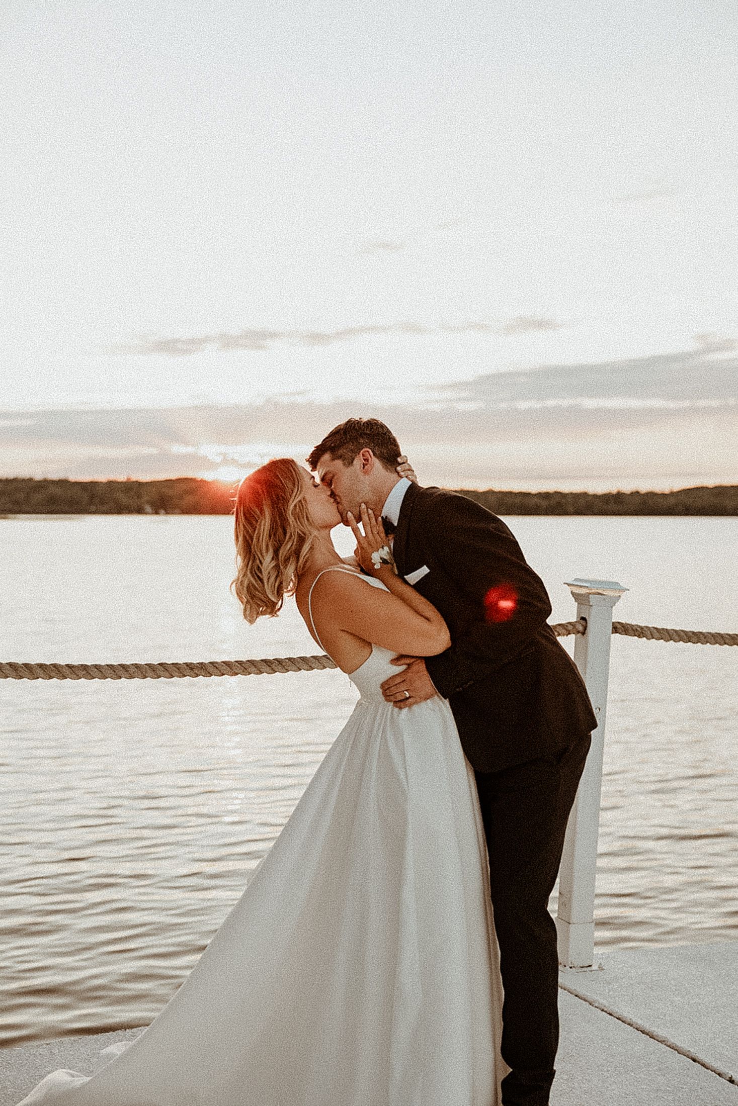A romantic couple in wedding attire share an intimate kiss on a boat dock at sunset over a serene lake.