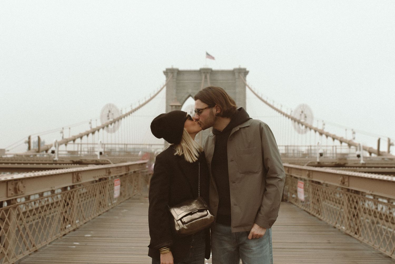 A couple in winter coats share an intimate moment on the Brooklyn Bridge while enjoying the sepia-toned cityscape.