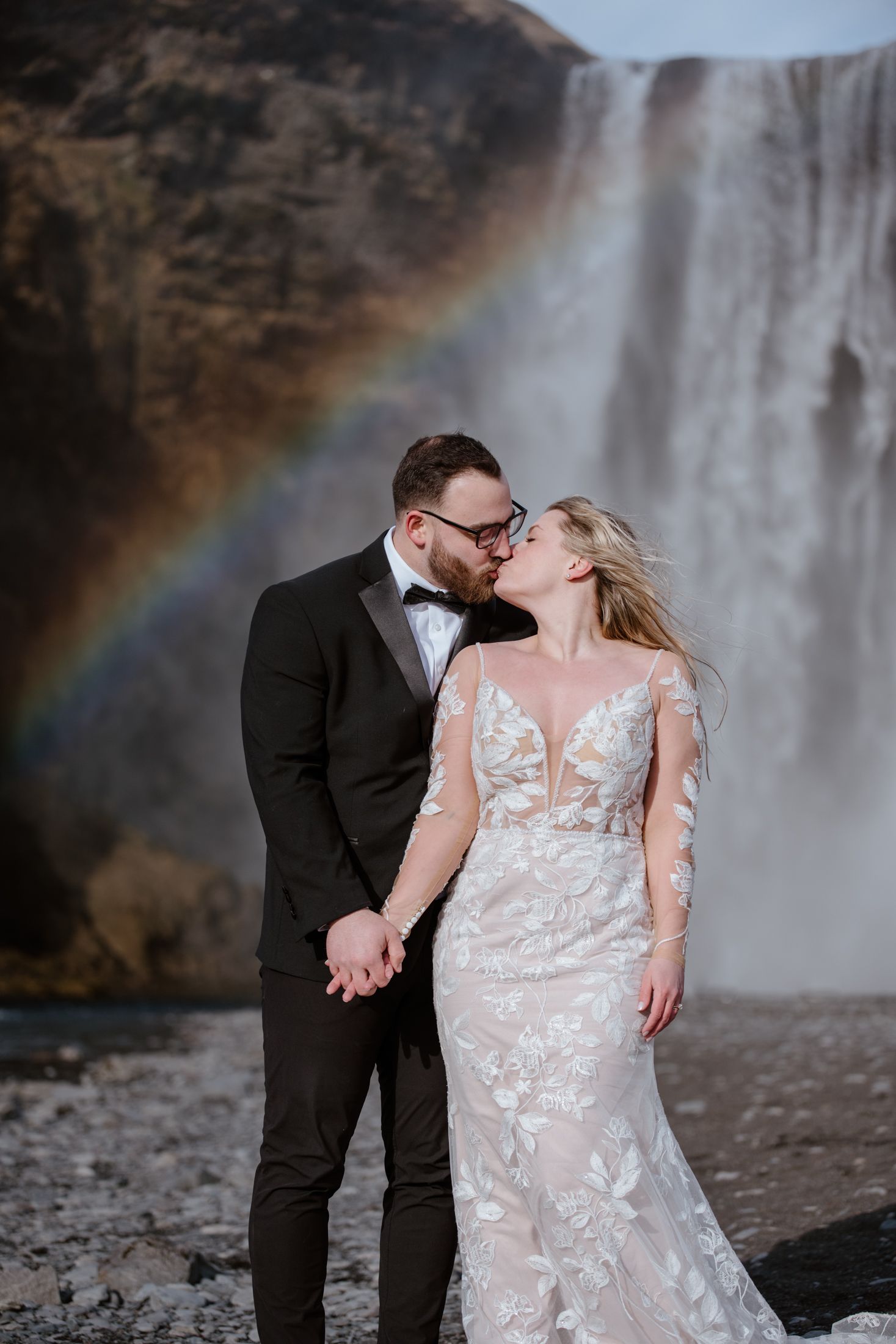 A romantic wedding couple embraces in front of a majestic waterfall with a vibrant rainbow in the background.