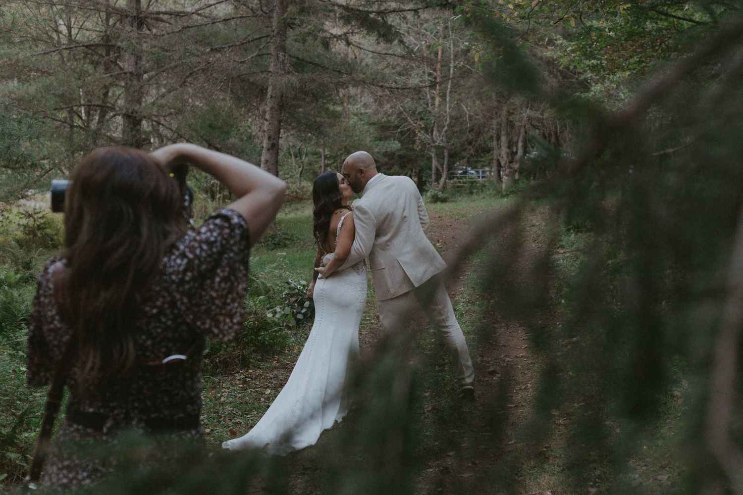 A photographer captures a couple in formal attire walking along a wooded path during an outdoor photo session.