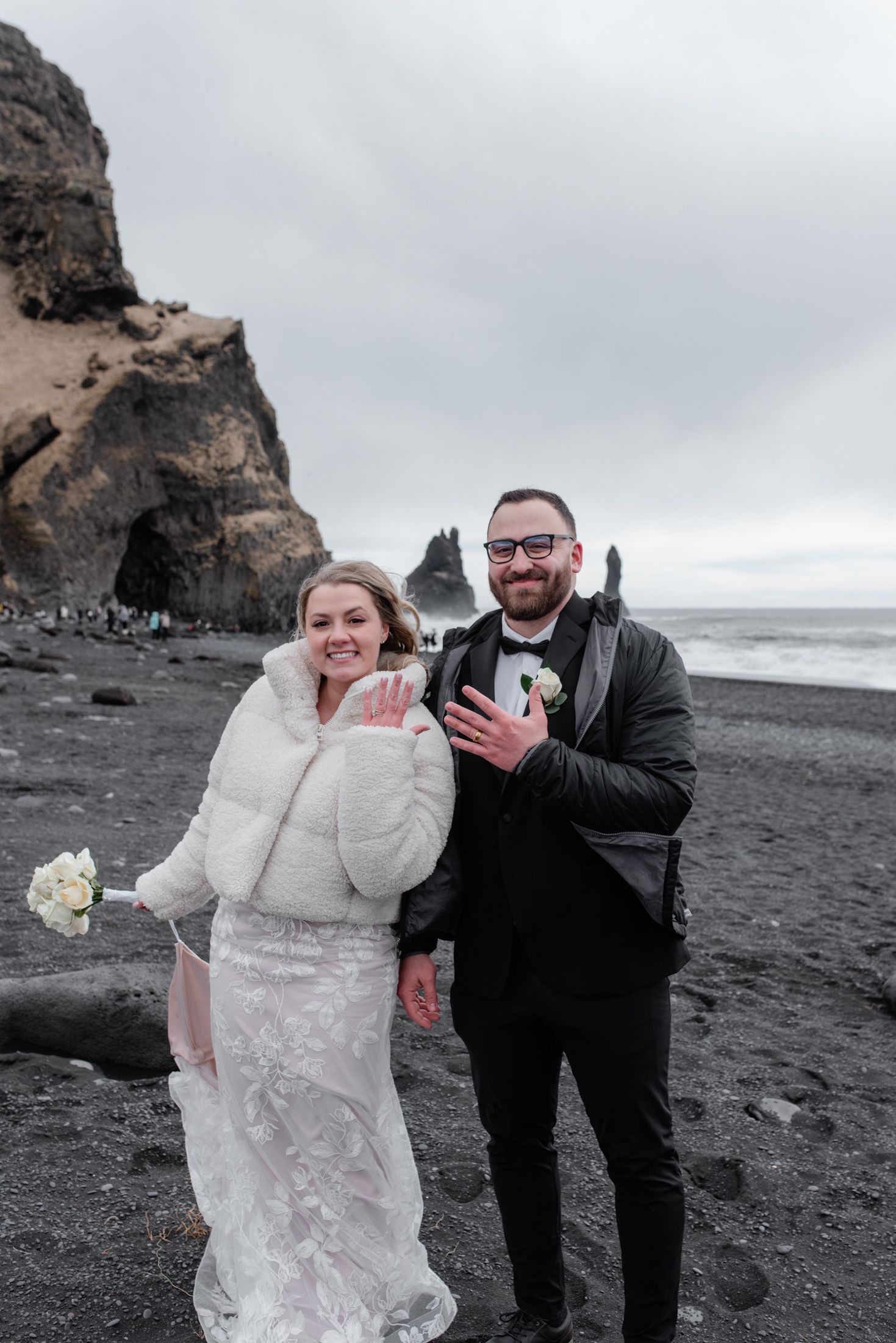 A couple in wedding attire pose together on a dramatic black sand beach with rocky cliffs and ocean waves in Iceland.