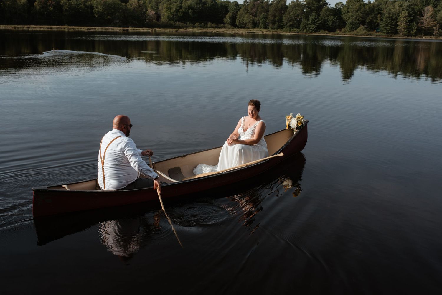 Three people in a wooden canoe glide across a still lake surrounded by pine trees on a tranquil day.