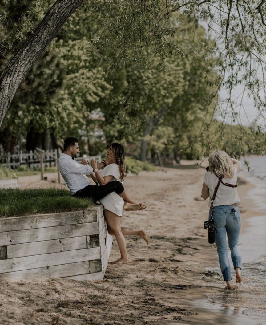 Two people enjoying a sunny day by a beach shoreline with trees and a wooden railing in the background.