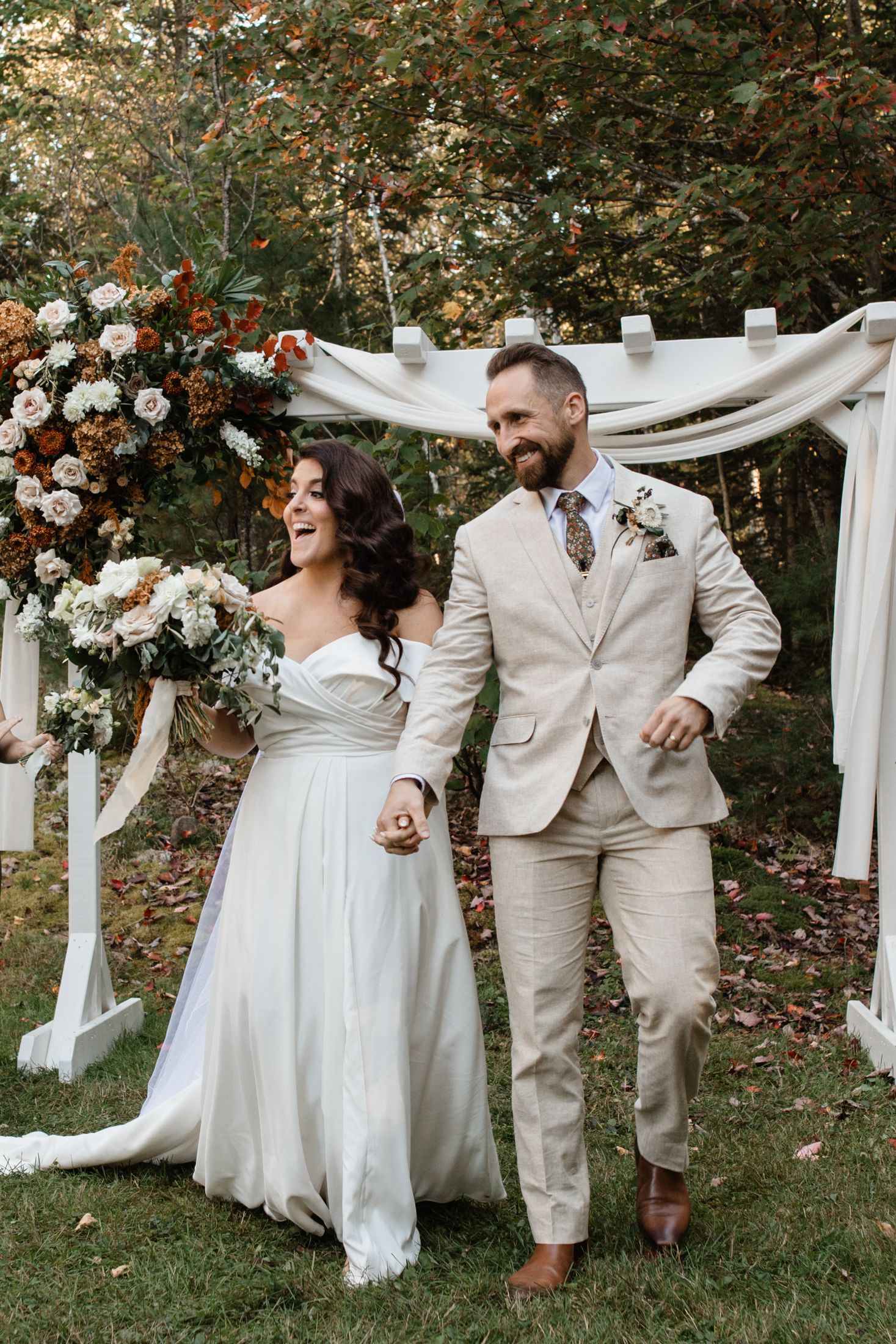 A joyful wedding couple laughs together under a white fabric arch decorated with flowers in an outdoor garden setting.