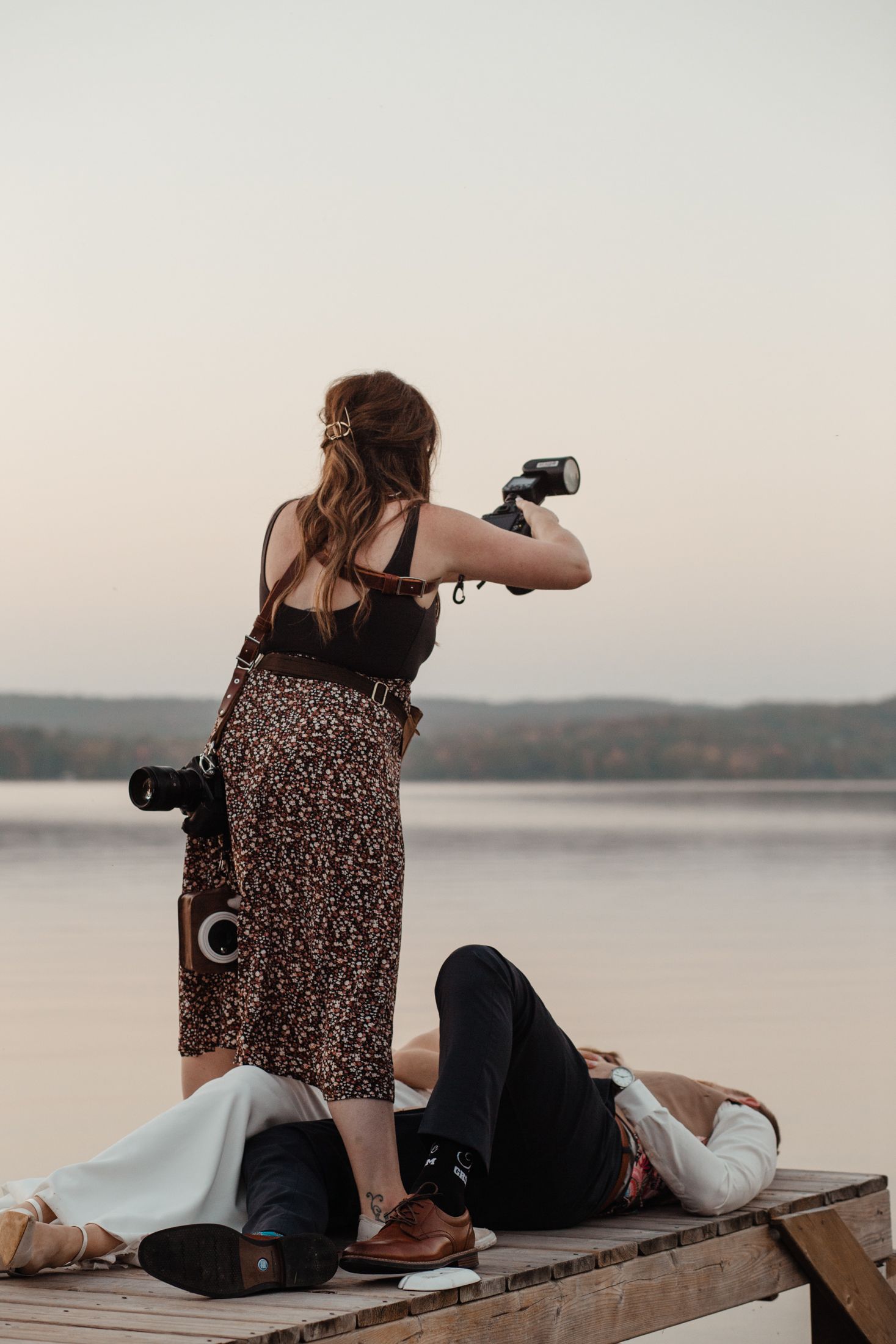 Woman in floral dress sitting on wooden deck with photography equipment overlooking lake at sunset.
