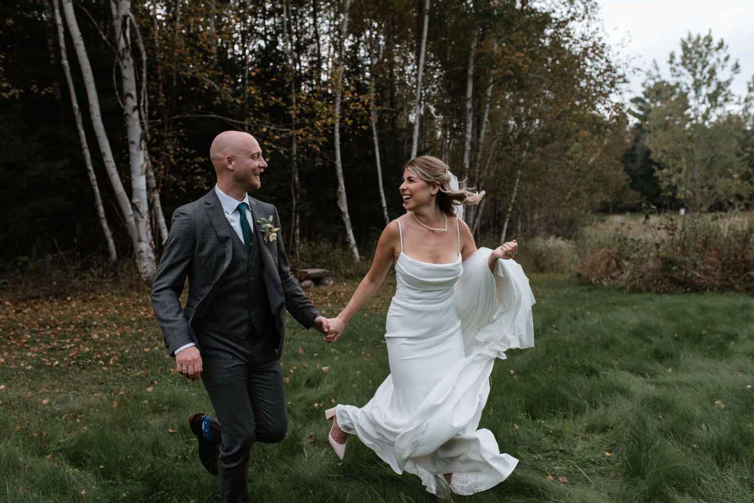 A bridal couple walks hand in hand through a grassy field near a wooded area on their wedding day.