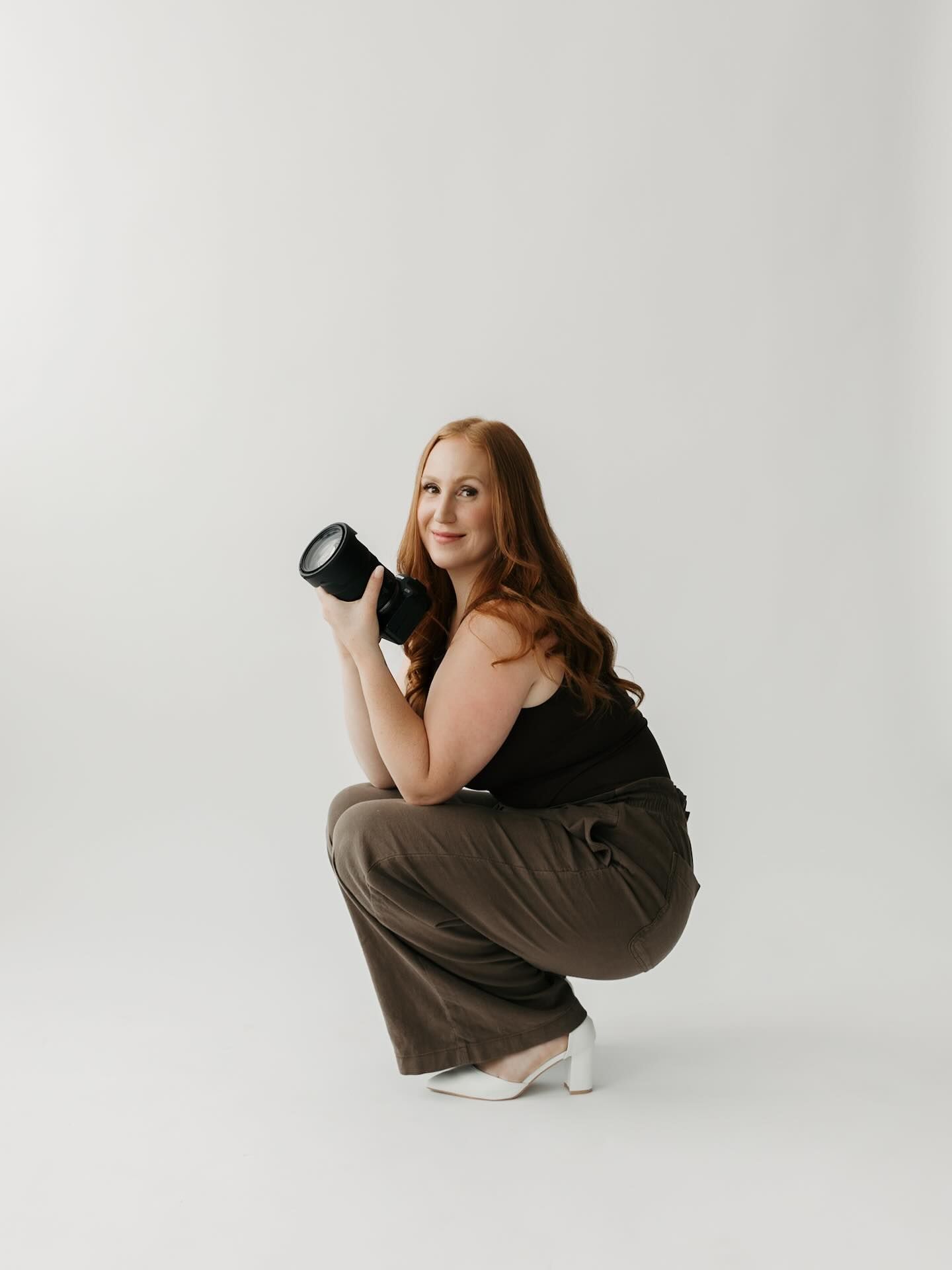 A photographer in a black top and brown pants crouches down while taking photos in a minimalist white studio setting.