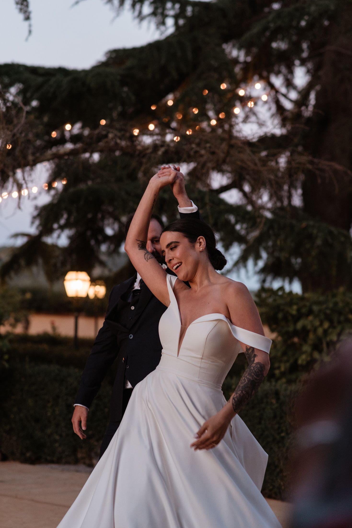 A couple dancing outdoors at twilight under string lights with bride wearing an off-shoulder white wedding dress.