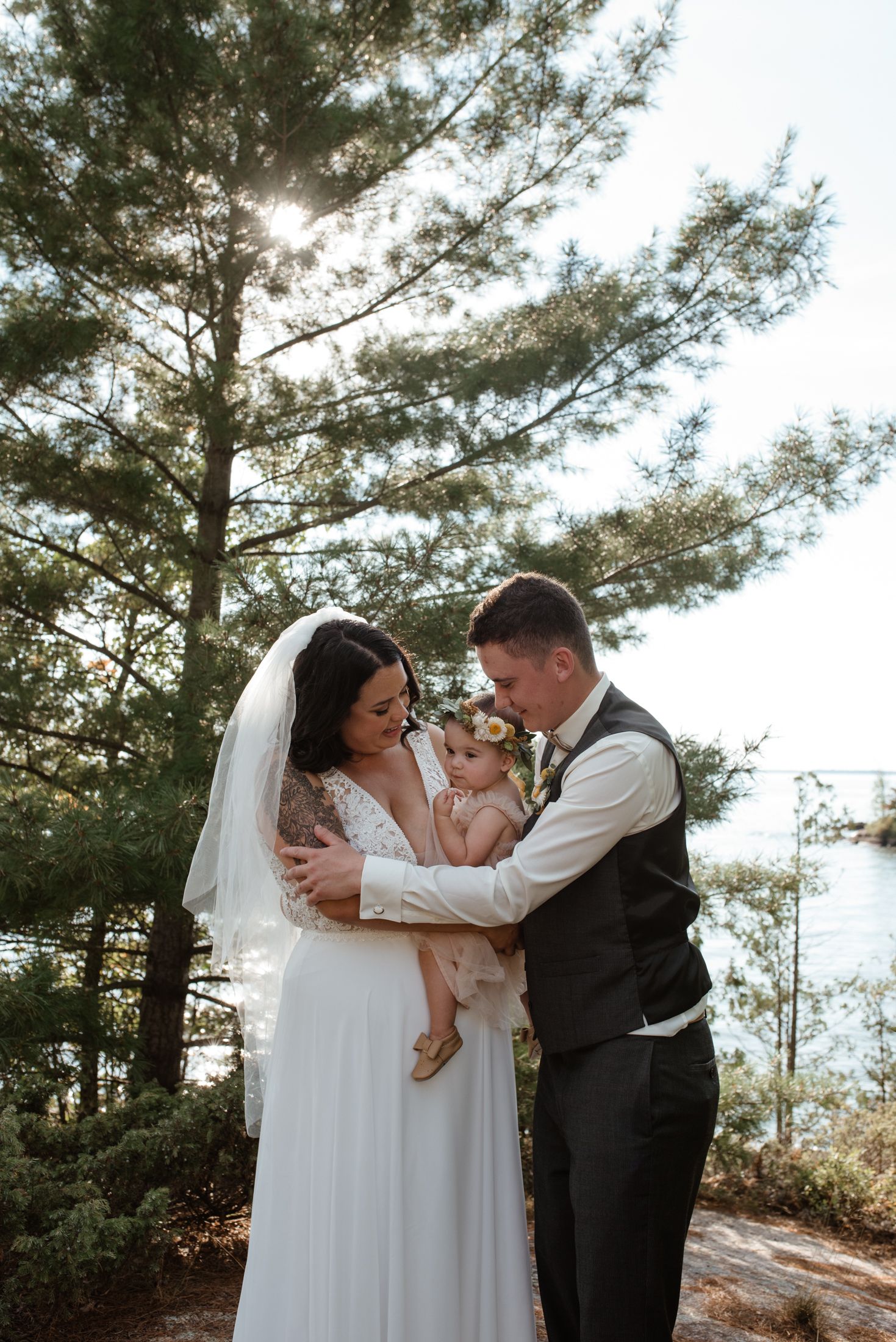 A couple shares a tender moment with a baby during their outdoor mountain wedding ceremony beneath towering pine trees.