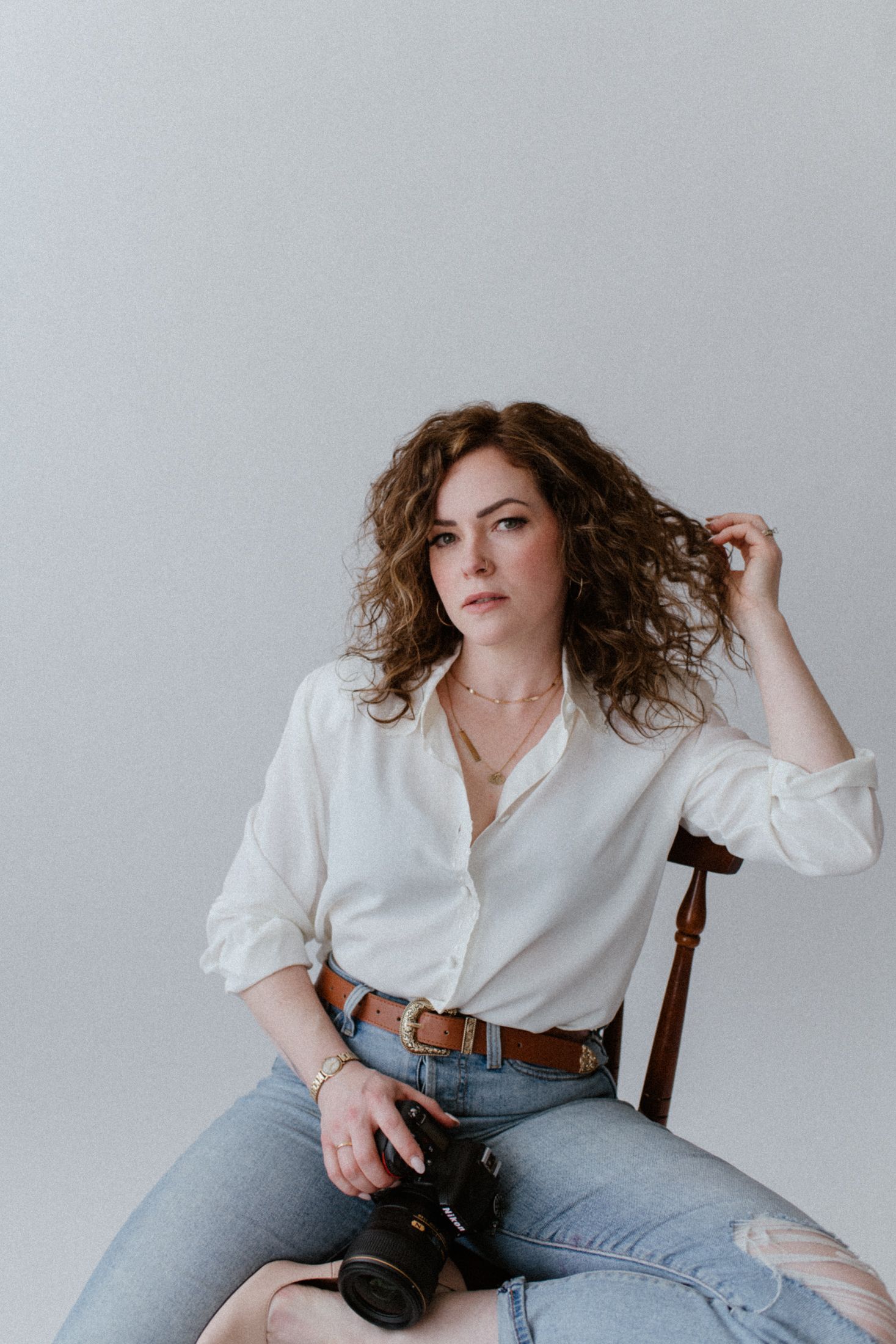 Photographer in casual attire with white button-down shirt and jeans sits holding a camera against neutral background.