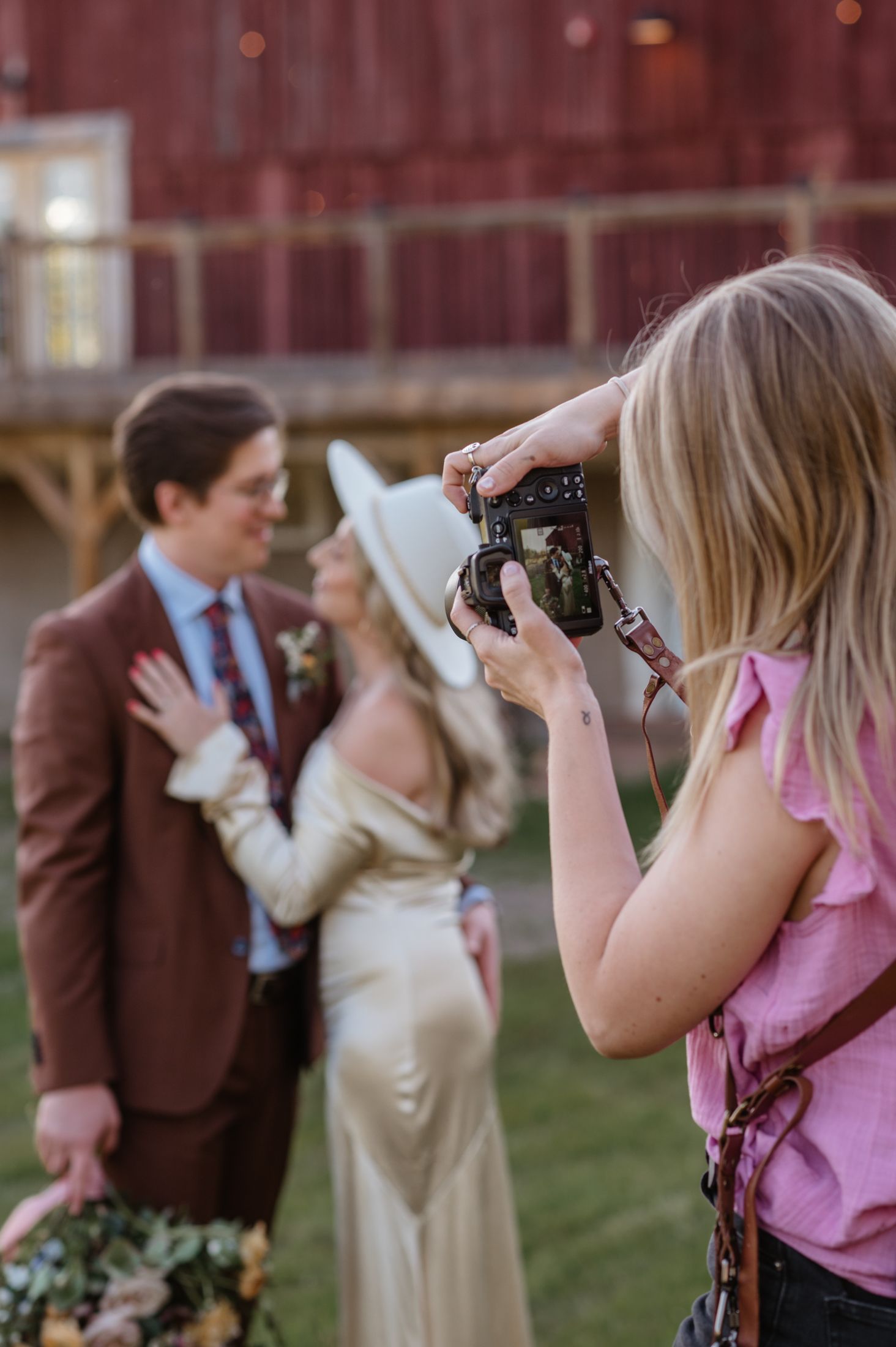 A photographer in a pink top captures a couple posing during a wedding photoshoot near a rustic barn setting.