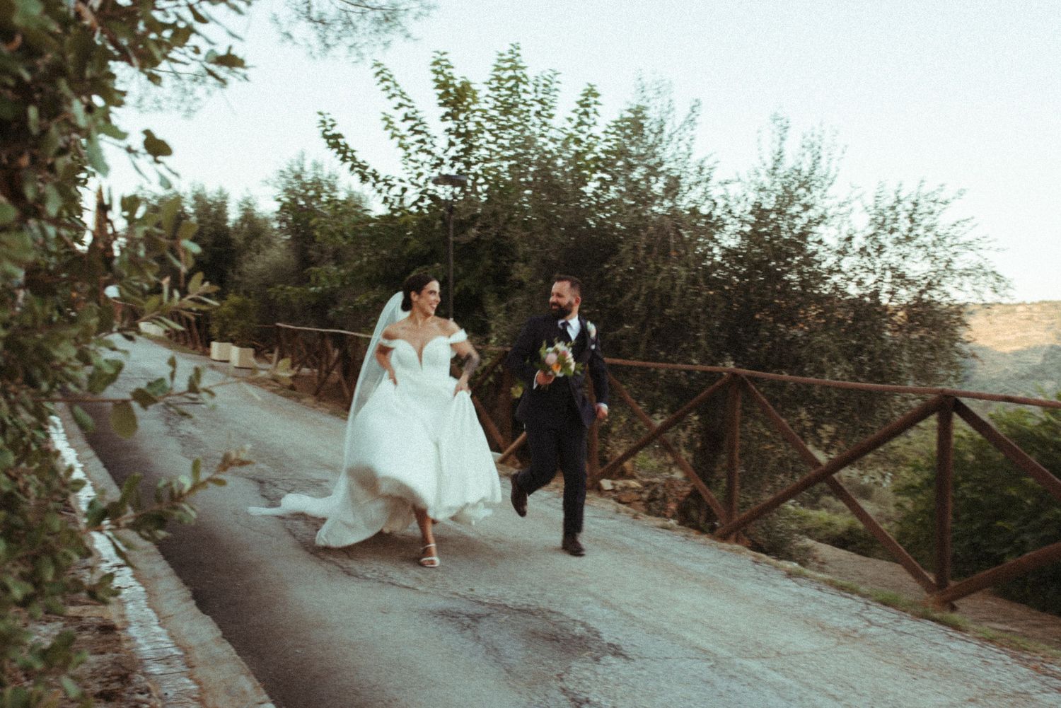 A couple in wedding attire runs playfully along a rural path with wooden fencing and lush greenery in the background.