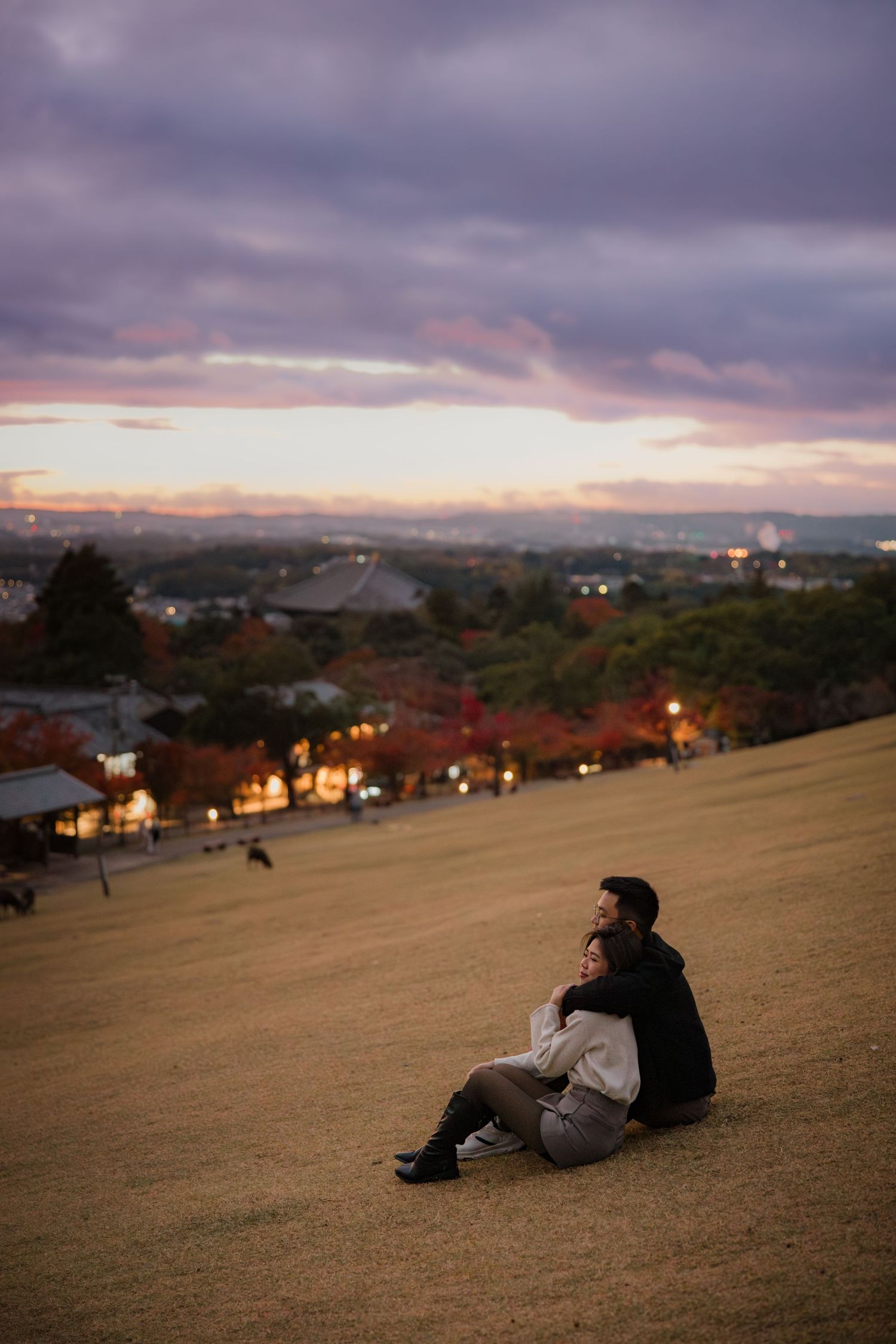 Person sitting on hillside overlooking city during sunset with dramatic purple sky.