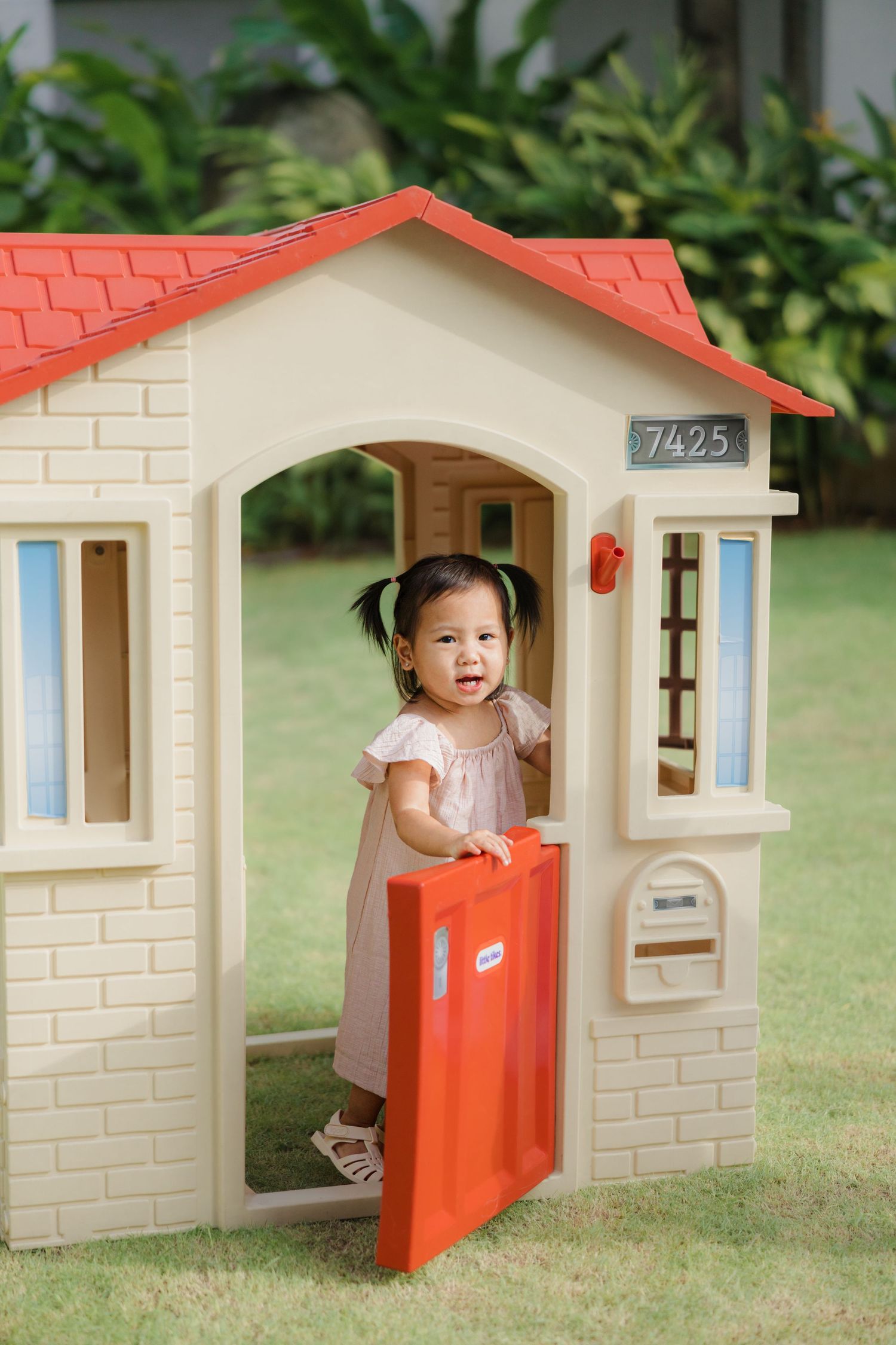 Small playhouse with red roof and cream walls stands in a garden with a child peeking through the orange door.