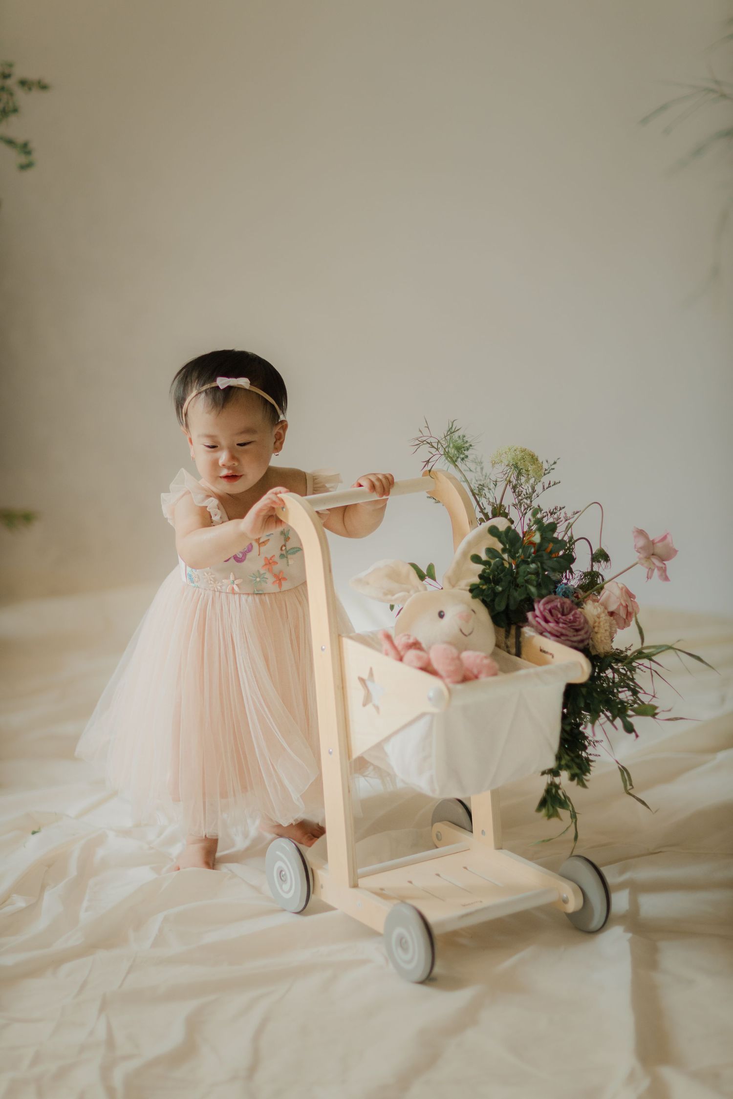 Child in pink tulle dress pushes wooden shopping cart with flowers and decorations.
